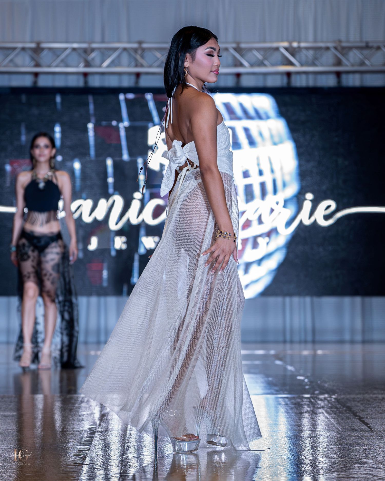 Flowing white sheer evening gown displayed on runway with dramatic backlighting.