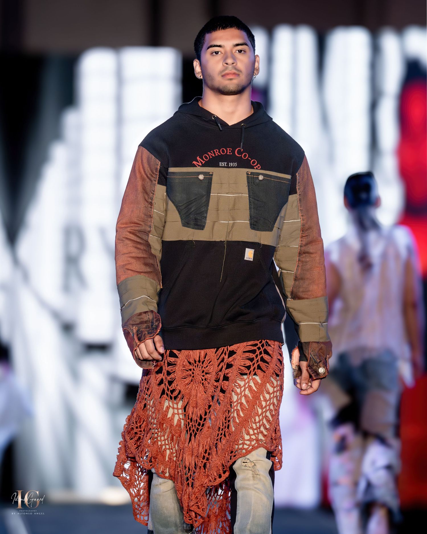Fashion model wearing workwear-inspired top and patterned brown skirt walks down illuminated runway.