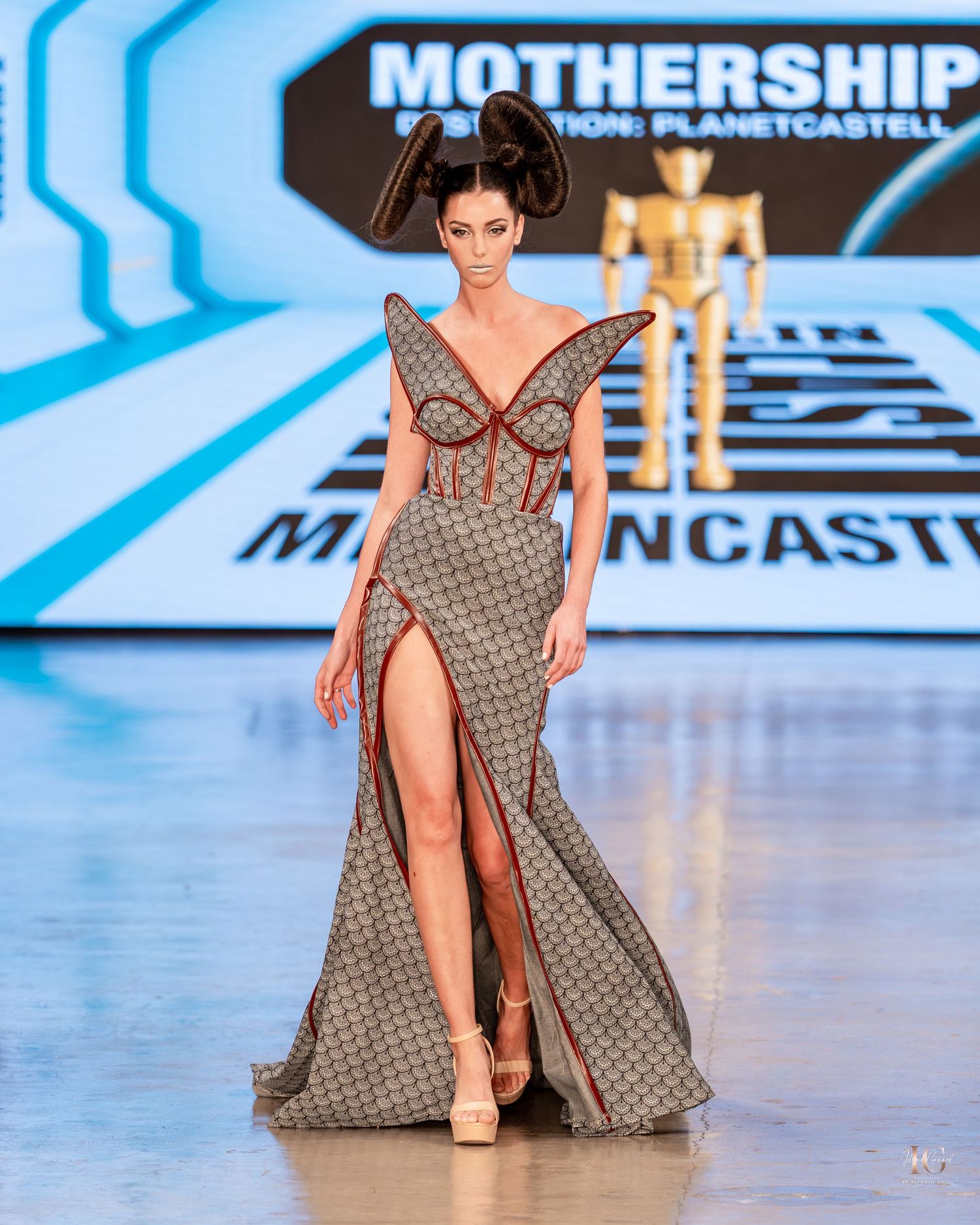 Model walks runway in gray geometric print dress with dramatic side slit at fashion show.
