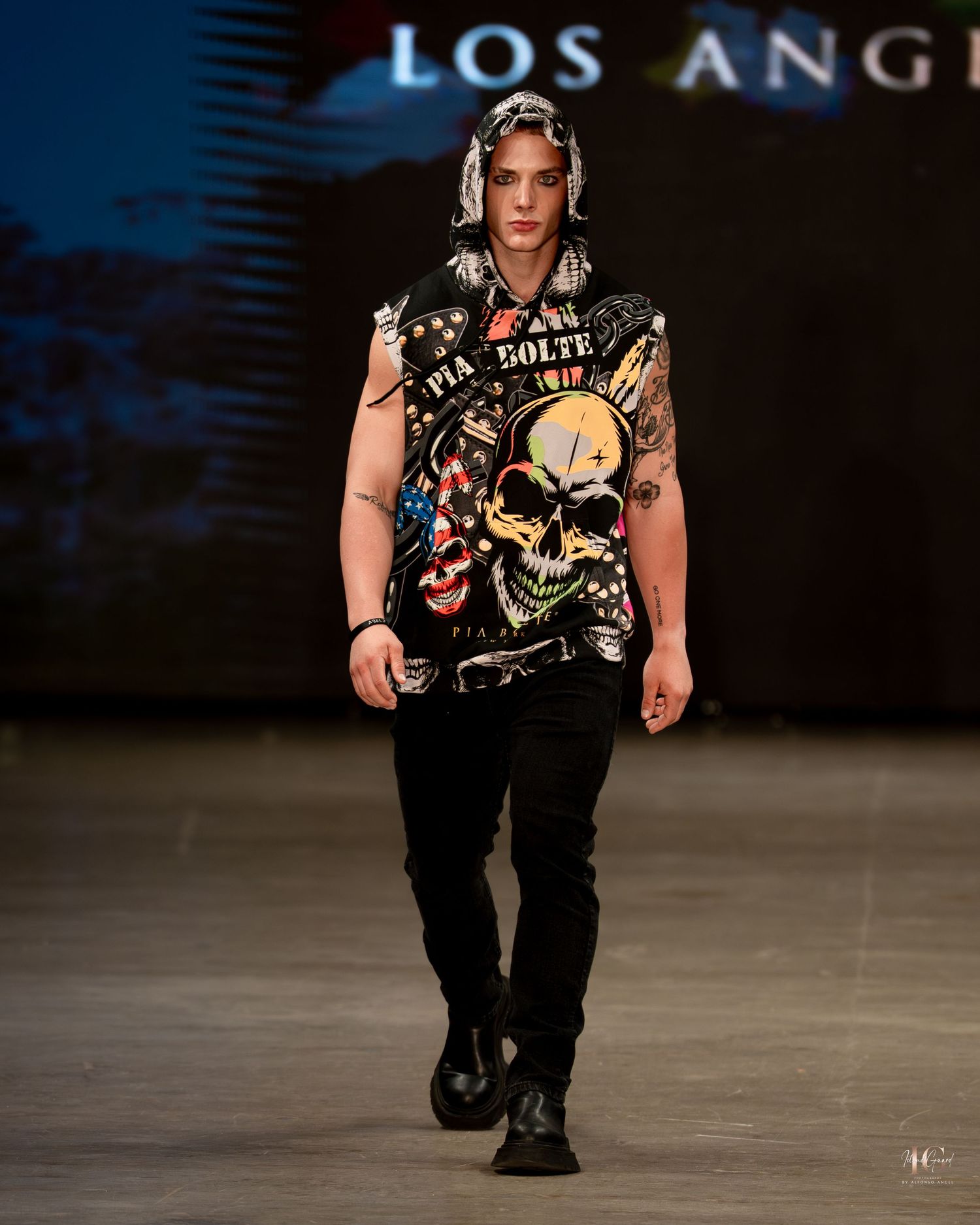 Model walks runway wearing black punk-inspired outfit with skull graphic t-shirt and combat boots during fashion show.