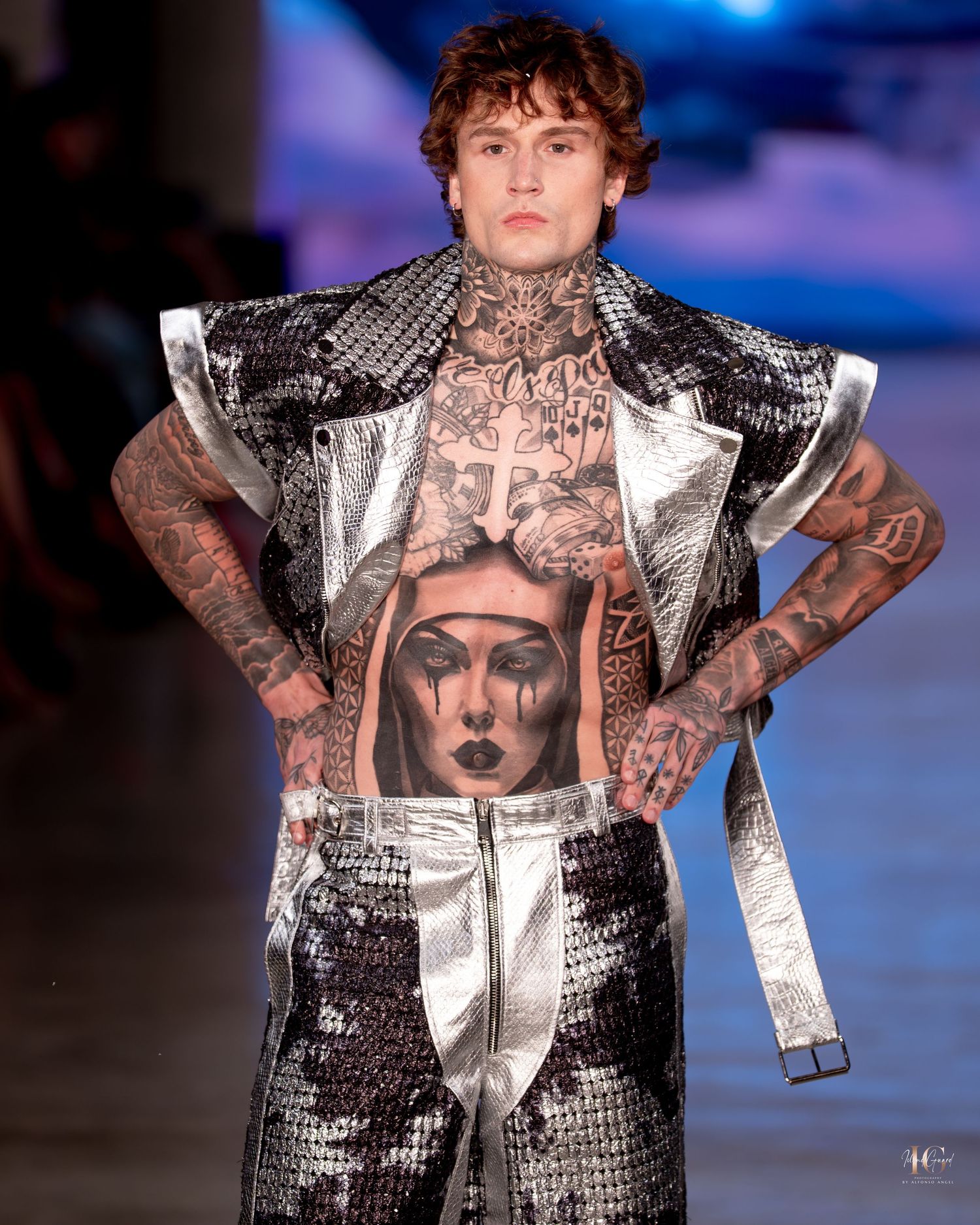 A fashion model walks the runway wearing a metallic sequined jacket with graphic print details in a dark setting.