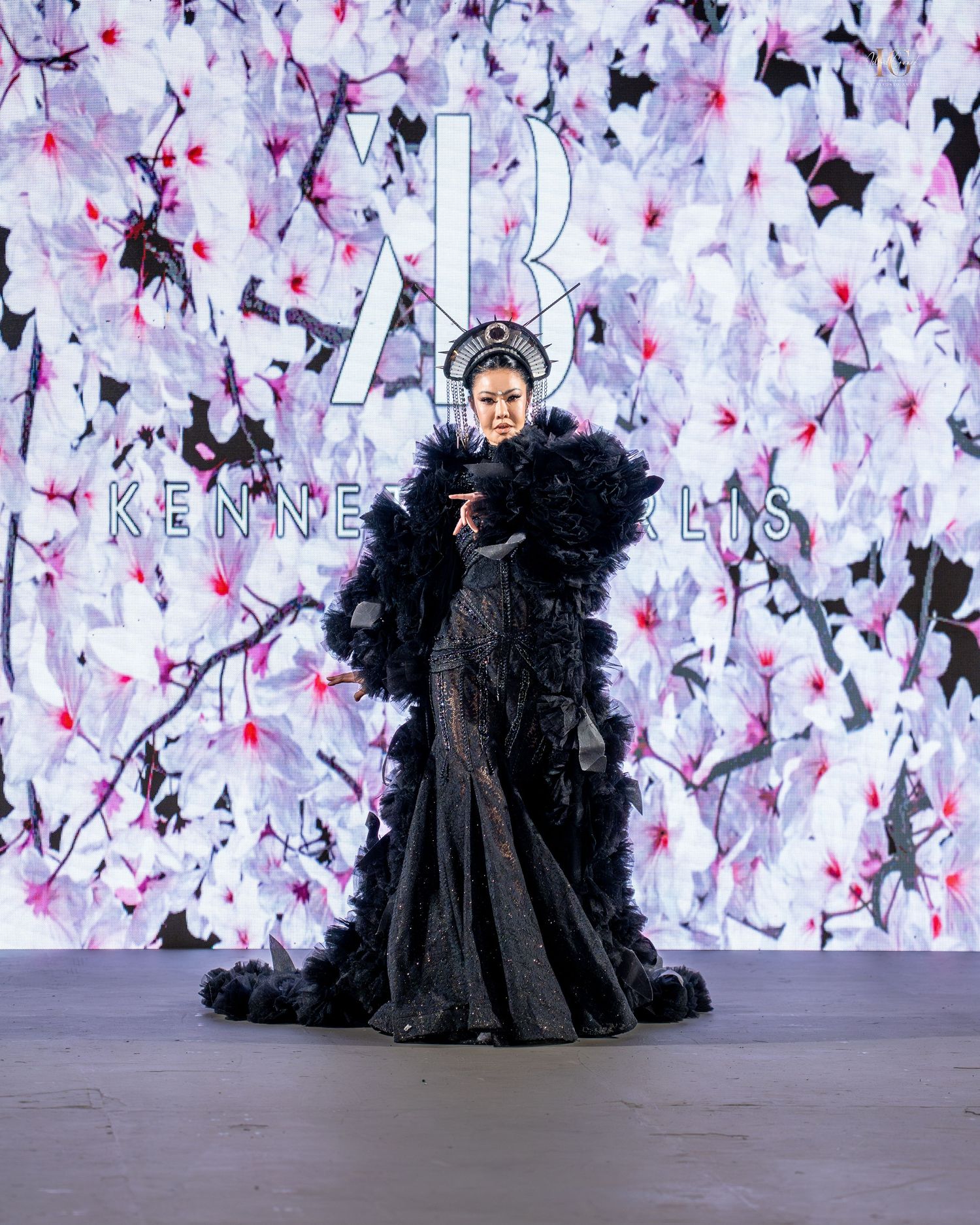 A model walks the runway in a dramatic black tulle gown with voluminous ruffles against a floral backdrop.