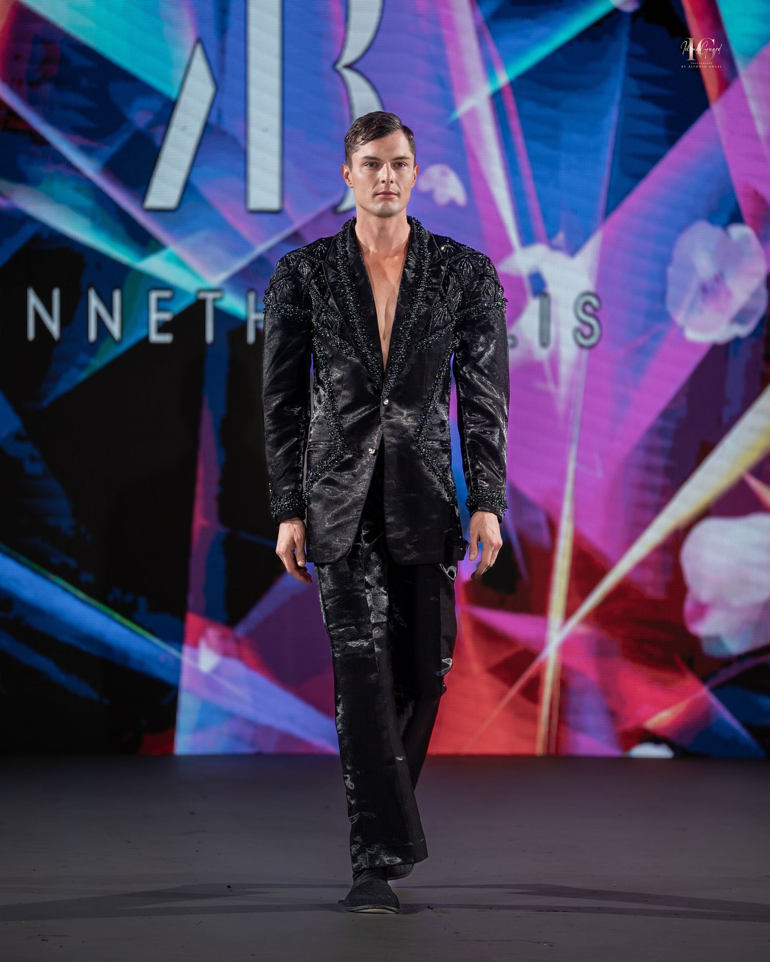 Model walks runway in black velvet suit during fashion show with colorful neon lighting effects in background.