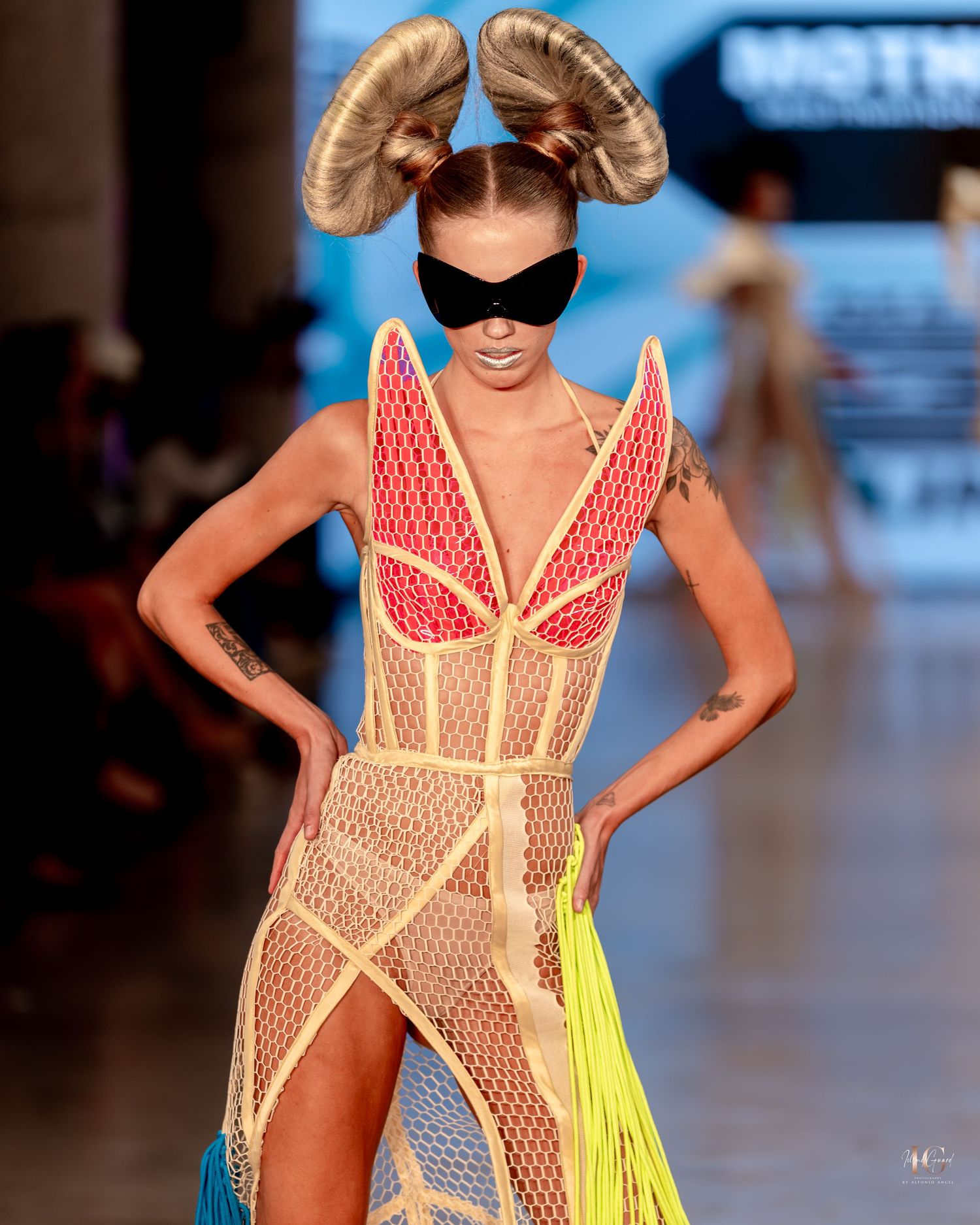 Model walks runway in sheer mesh dress with red geometric details, dramatic ram horn hairstyle and black eye mask at fashion show.