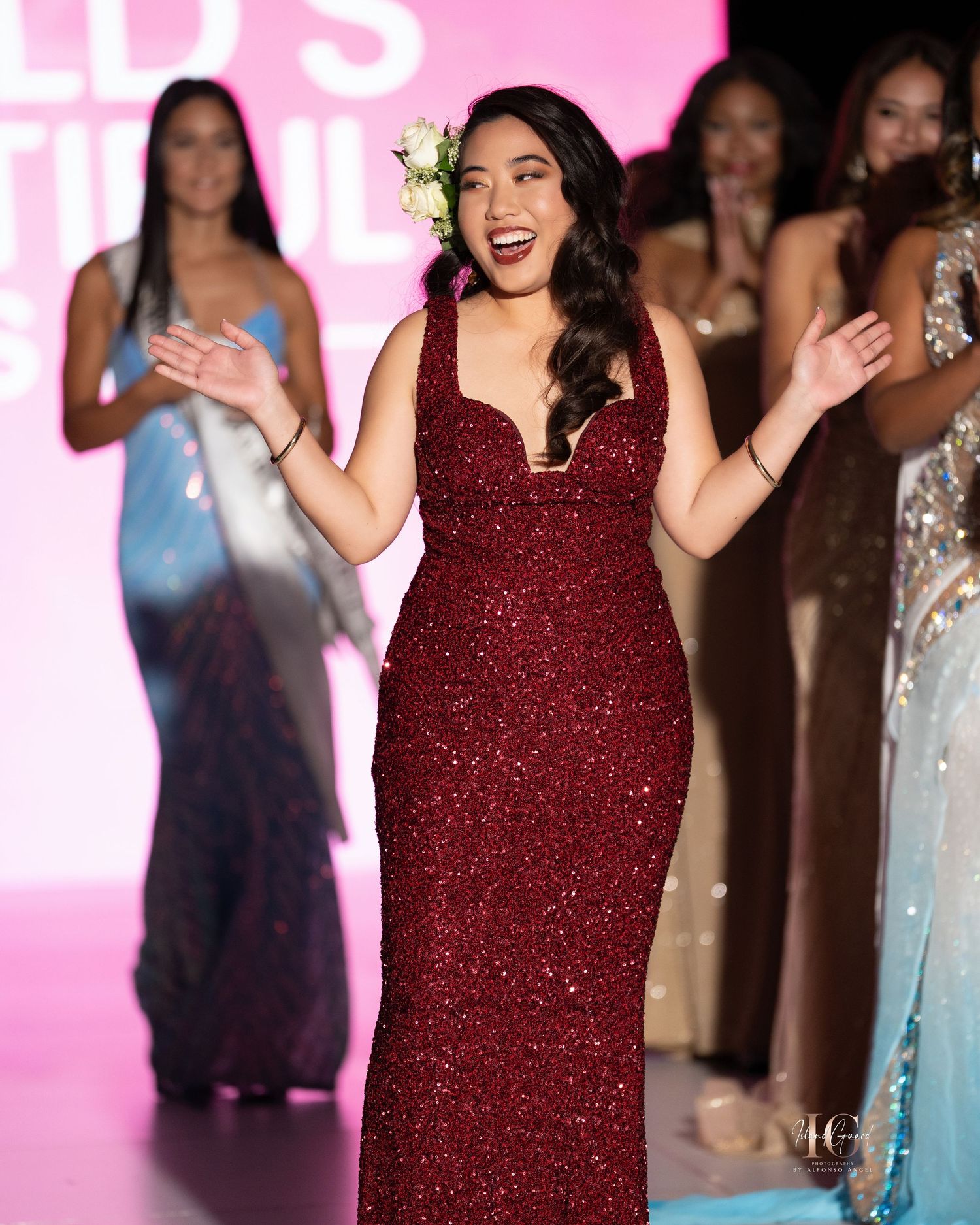 A model walks the runway in a sparkling burgundy evening gown at a fashion show with pink lighting.