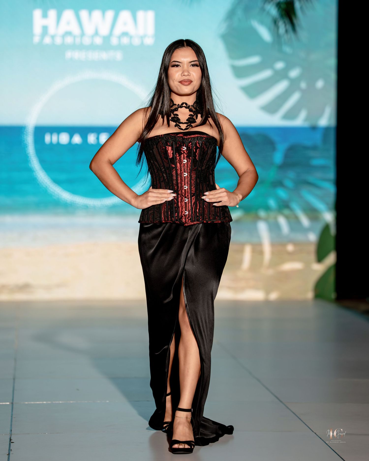 Model walks runway in black evening gown with corset detail at tropical-themed fashion show.
