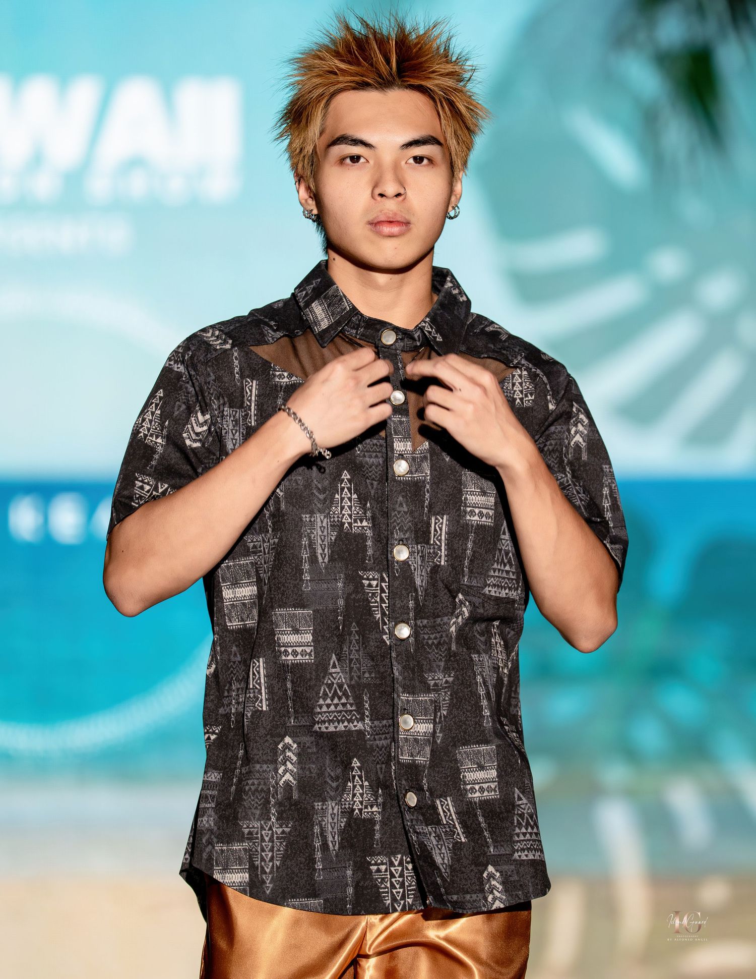 Model in a black patterned short-sleeve button-up shirt poses on runway against blue backdrop.