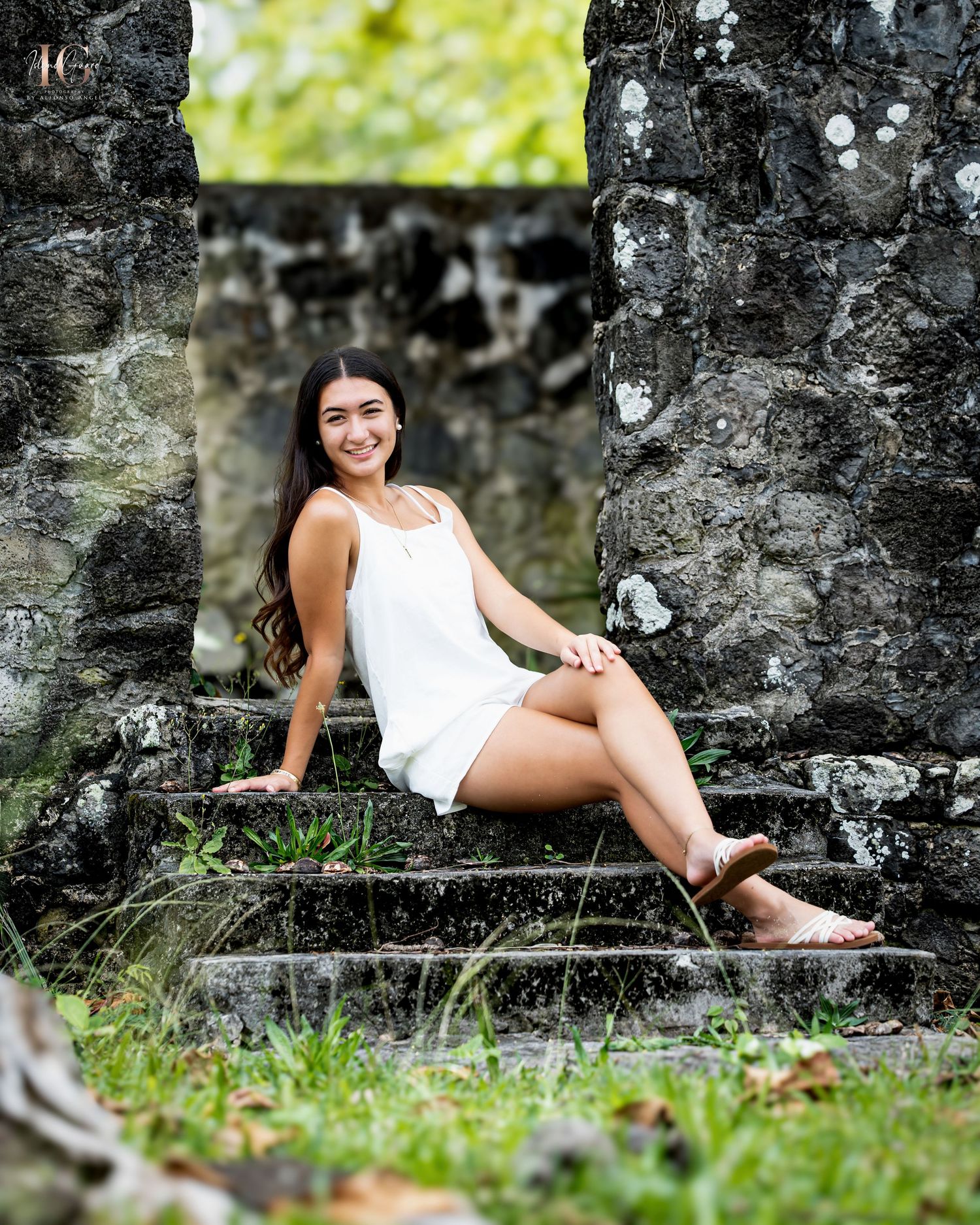 Portrait against stone ruins with sunlit greenery in the background showing someone in a white summer dress seated on steps.