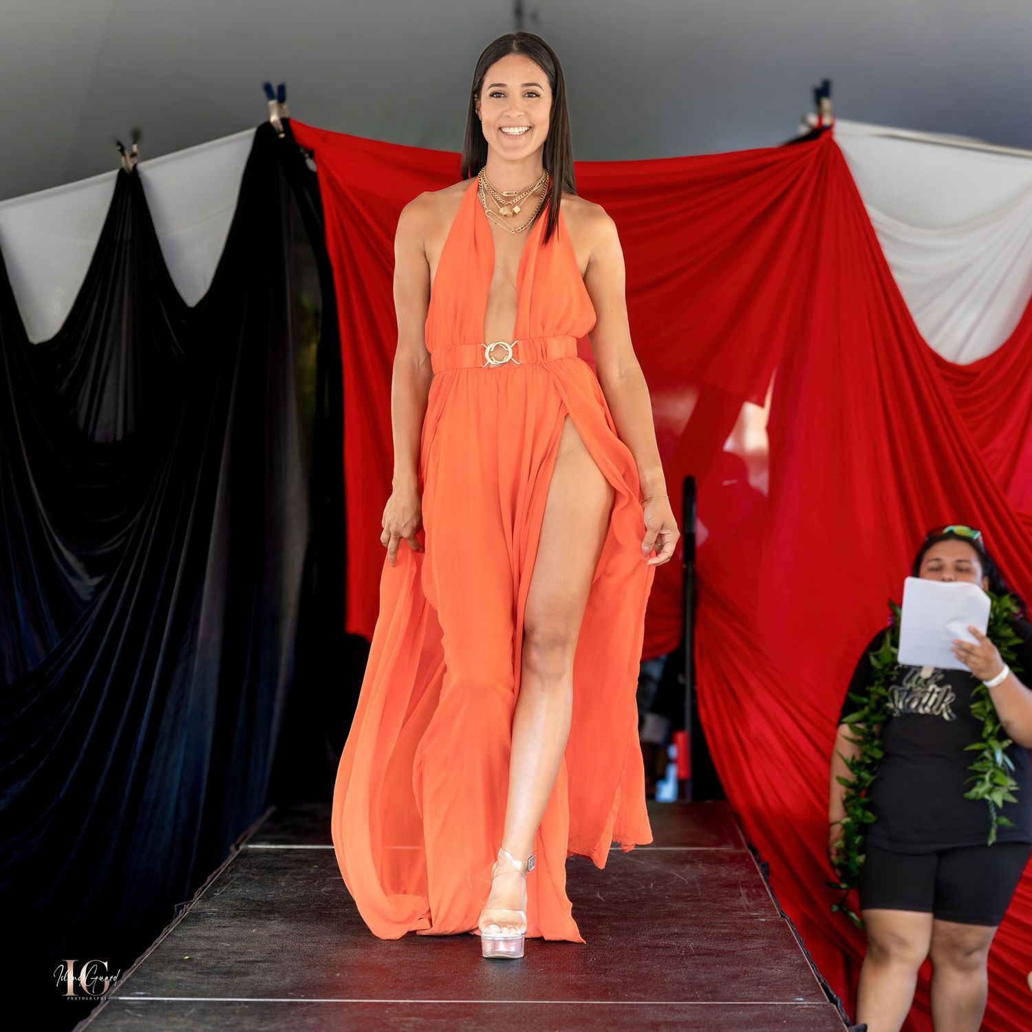Model walks runway in flowing coral maxi dress with thigh-high slit against dramatic backdrop.