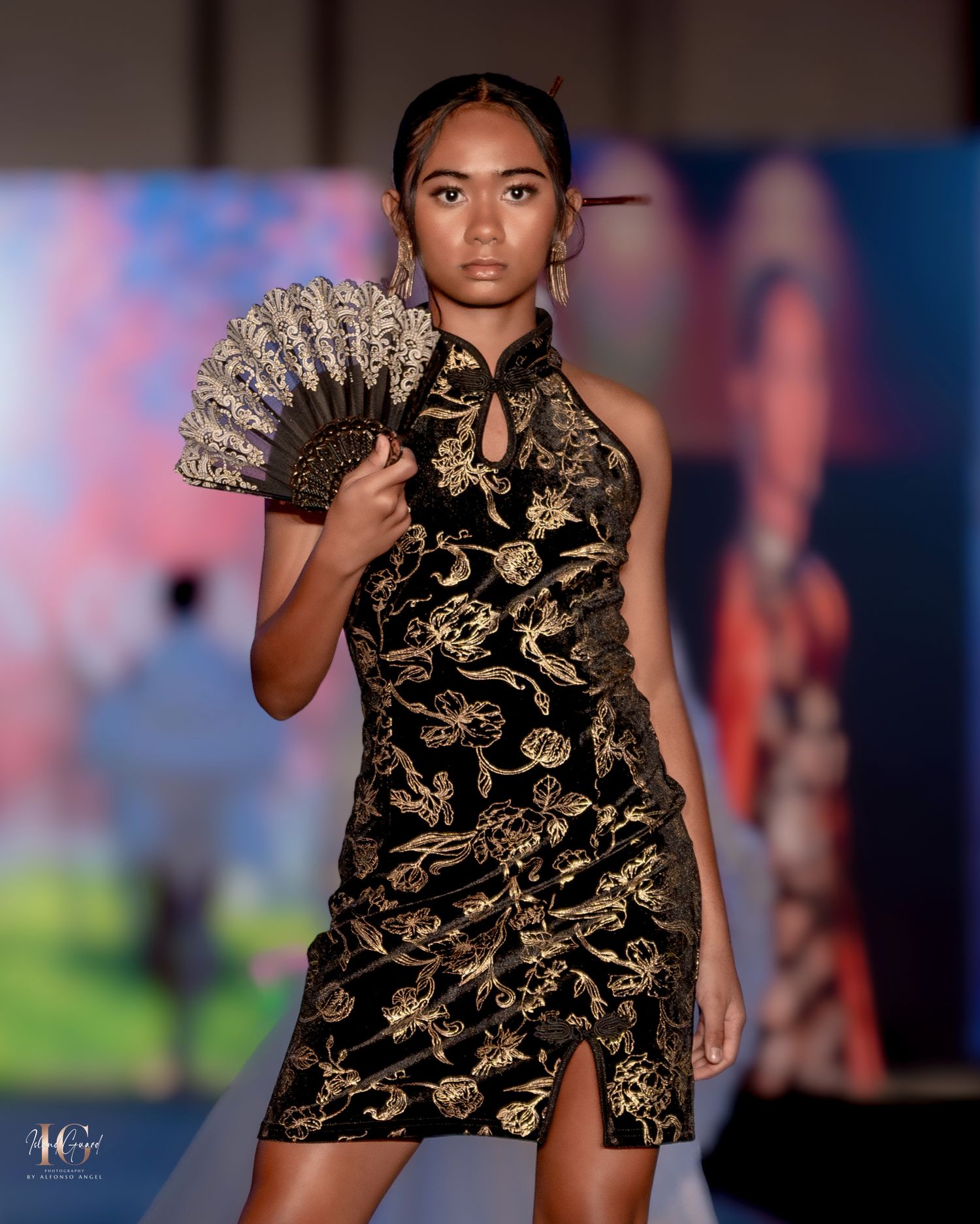 Fashion model walks runway in black and gold patterned designer dress during fashion show.