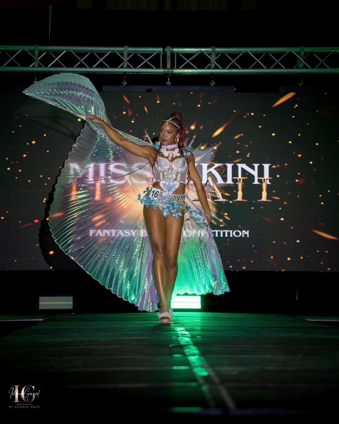 A glowing performer wearing an illuminated fantasy costume with flowing green wings creates a dramatic silhouette on stage.
