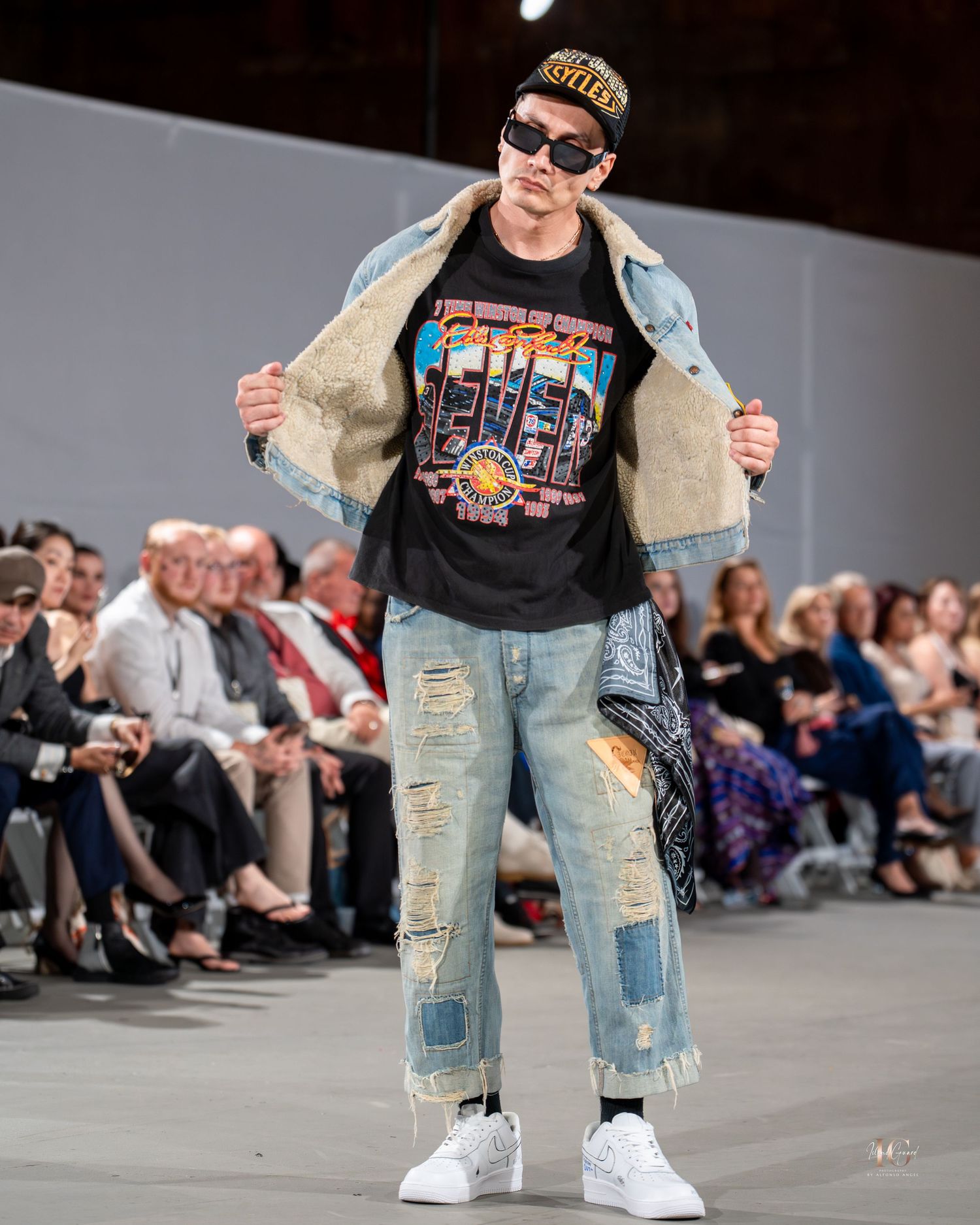 Model walks runway in distressed denim, graphic t-shirt and denim jacket at fashion show with audience in background.