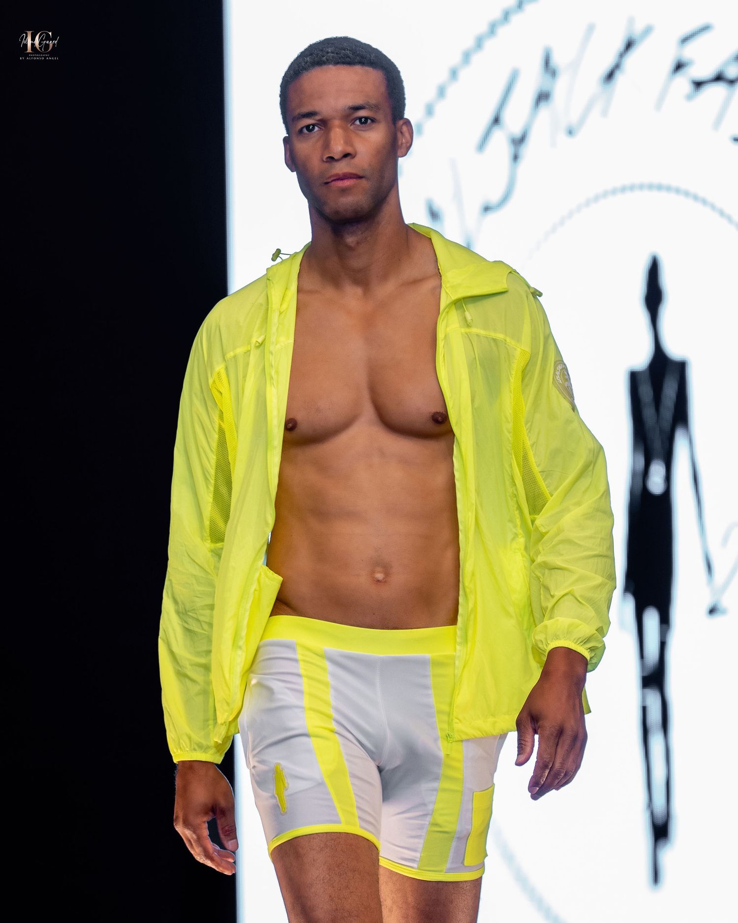 Model walks runway wearing neon yellow windbreaker jacket and white shorts with yellow accents during fashion show.