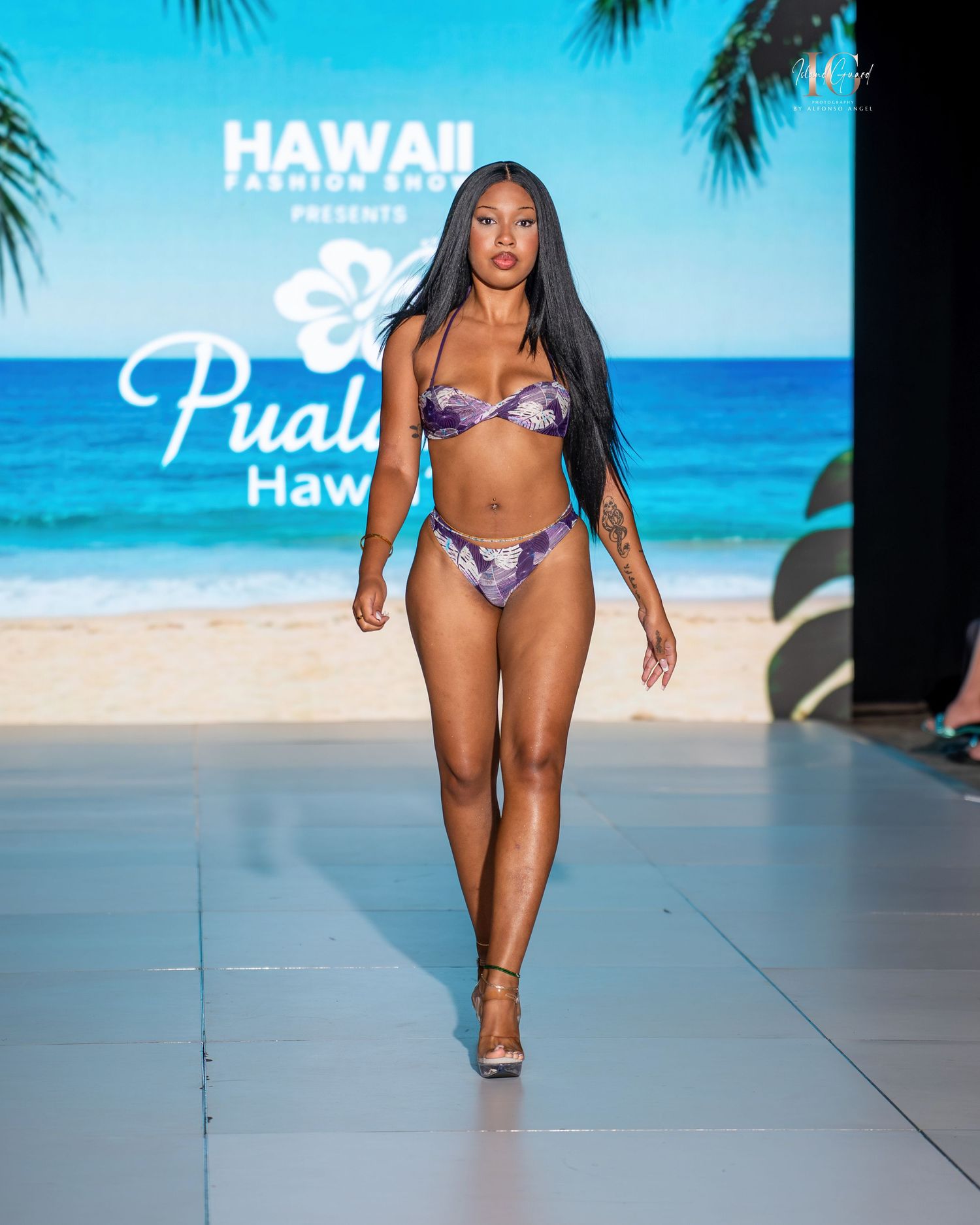Models walk the runway in tropical print swimwear at a Hawaii fashion show on a beachfront catwalk.