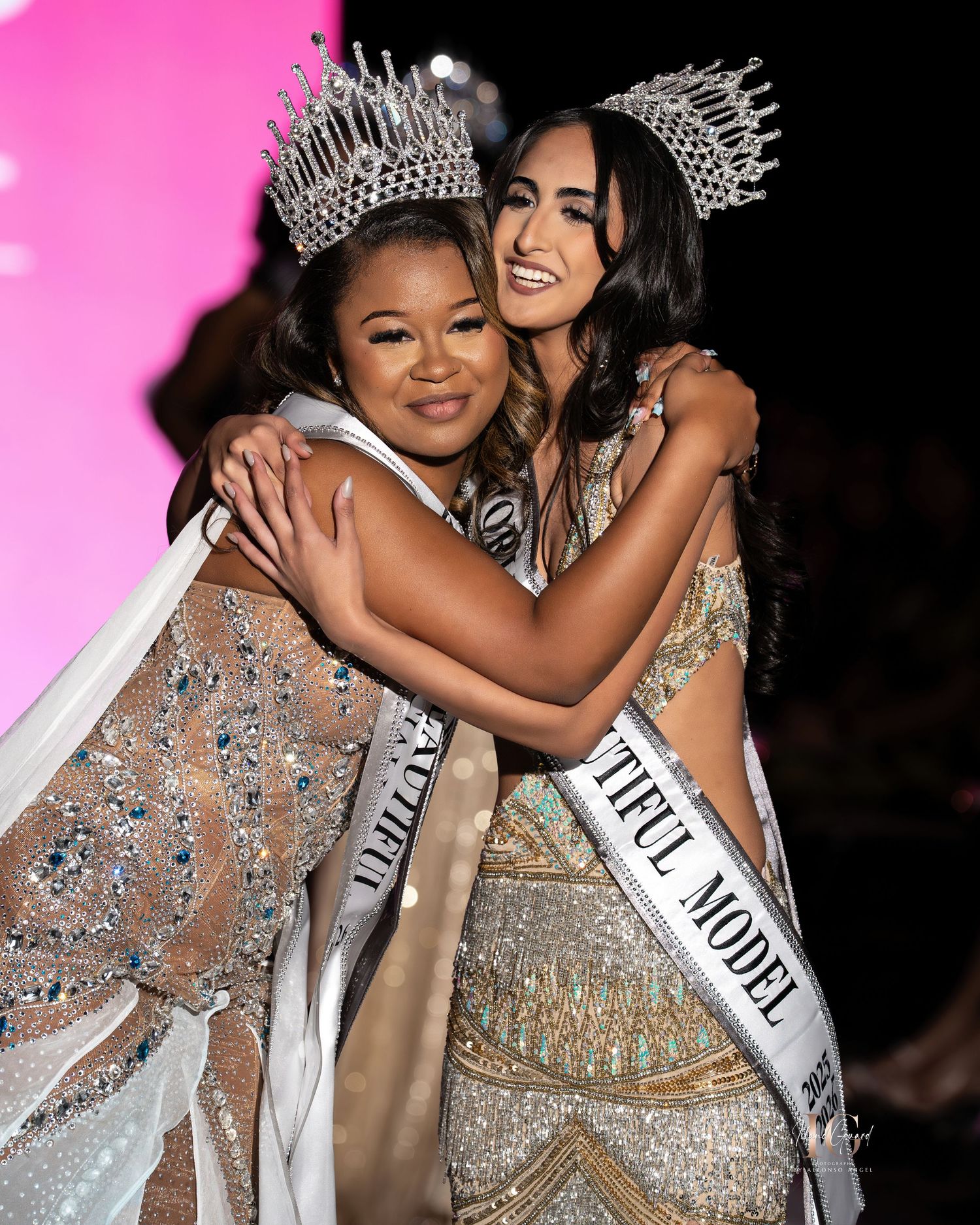 Pageant contestants wearing sparkling evening gowns and crowns embrace on stage with pink backdrop.