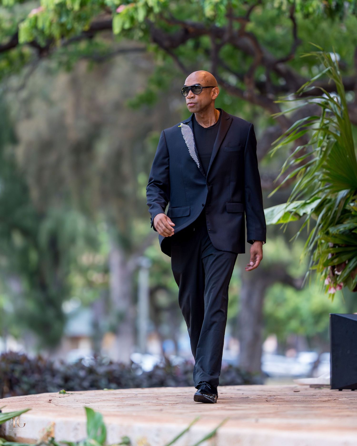 Someone in a dark suit walks along a path in a lush garden setting with trees and greenery in the background.