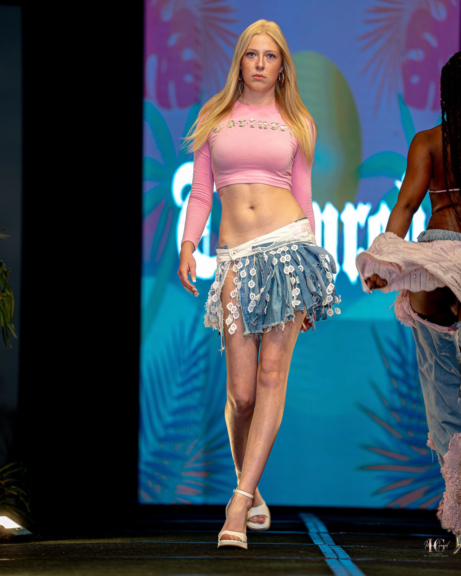 Fashion model walks runway in pink crop top and floral denim shorts with colorful stage lighting in background.