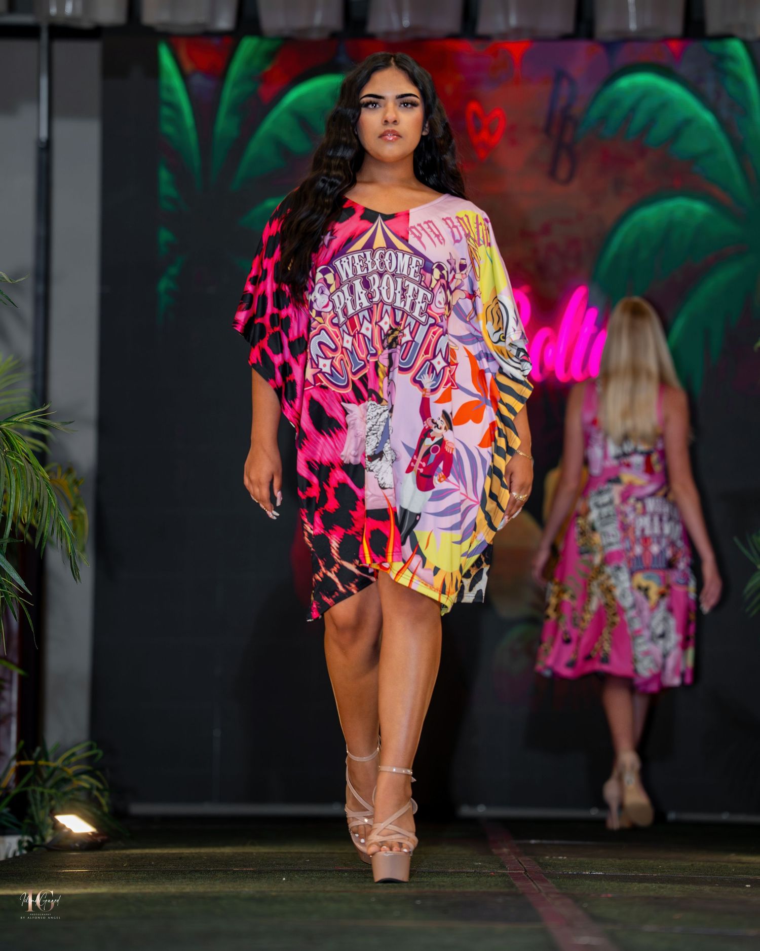 A fashion runway show features models walking in vibrant pink and graffiti-style printed outfits against a dark backdrop.