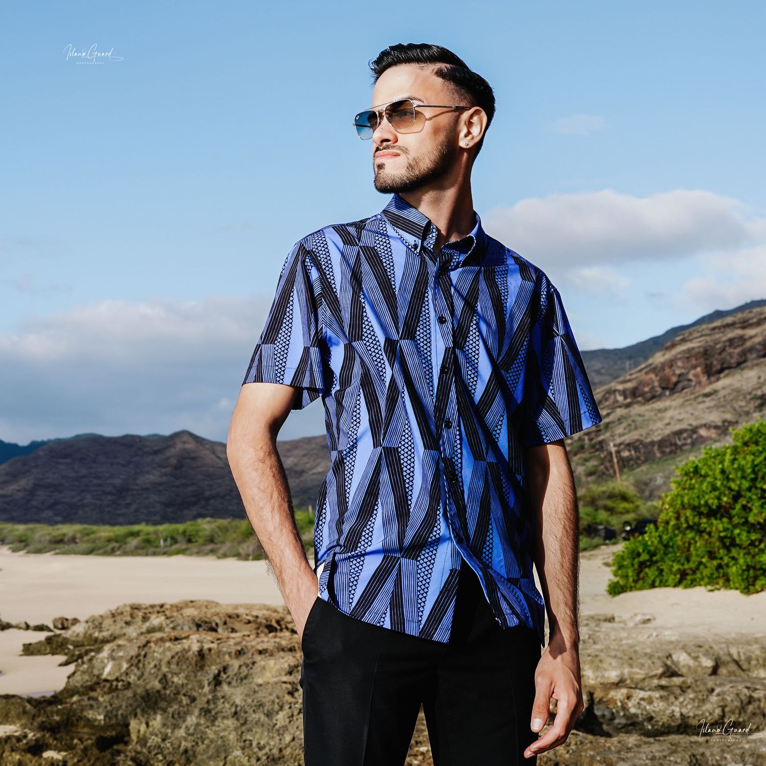 Man wearing blue geometric pattern shirt poses against mountainous landscape on bright sunny day.