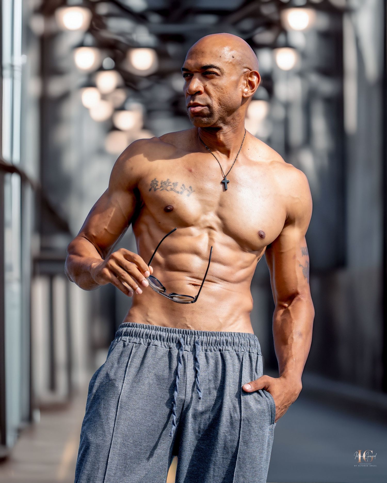 Muscular shirtless athlete performs torso twisting exercises in a gray sweatpants along an industrial corridor.