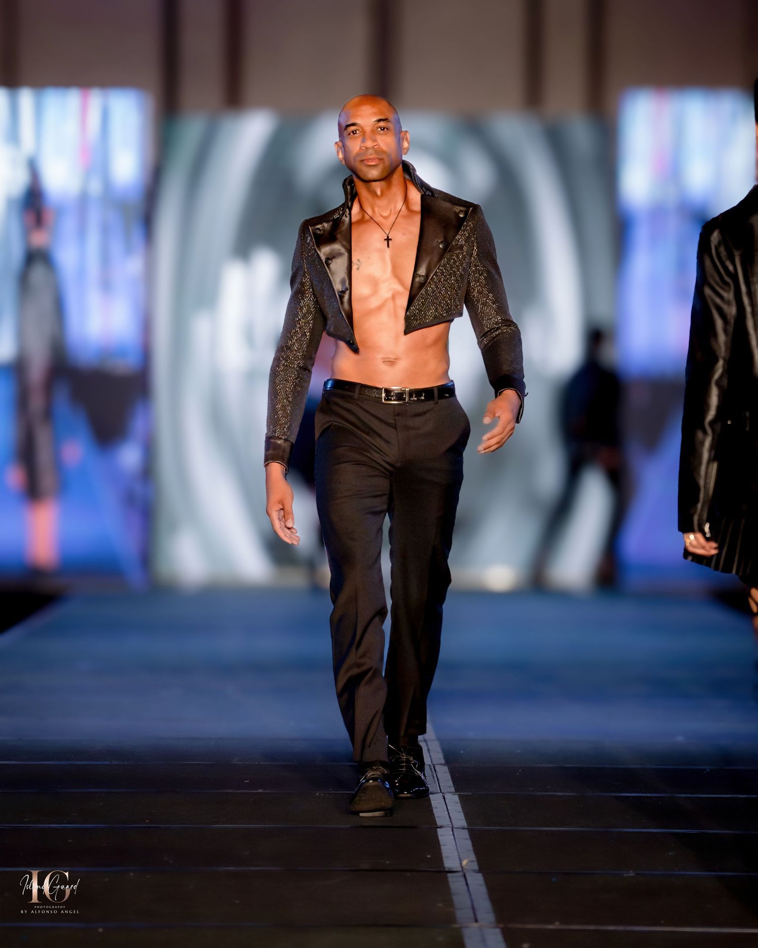 Model walks runway in sleek black leather blazer and fitted pants against illuminated backdrop.