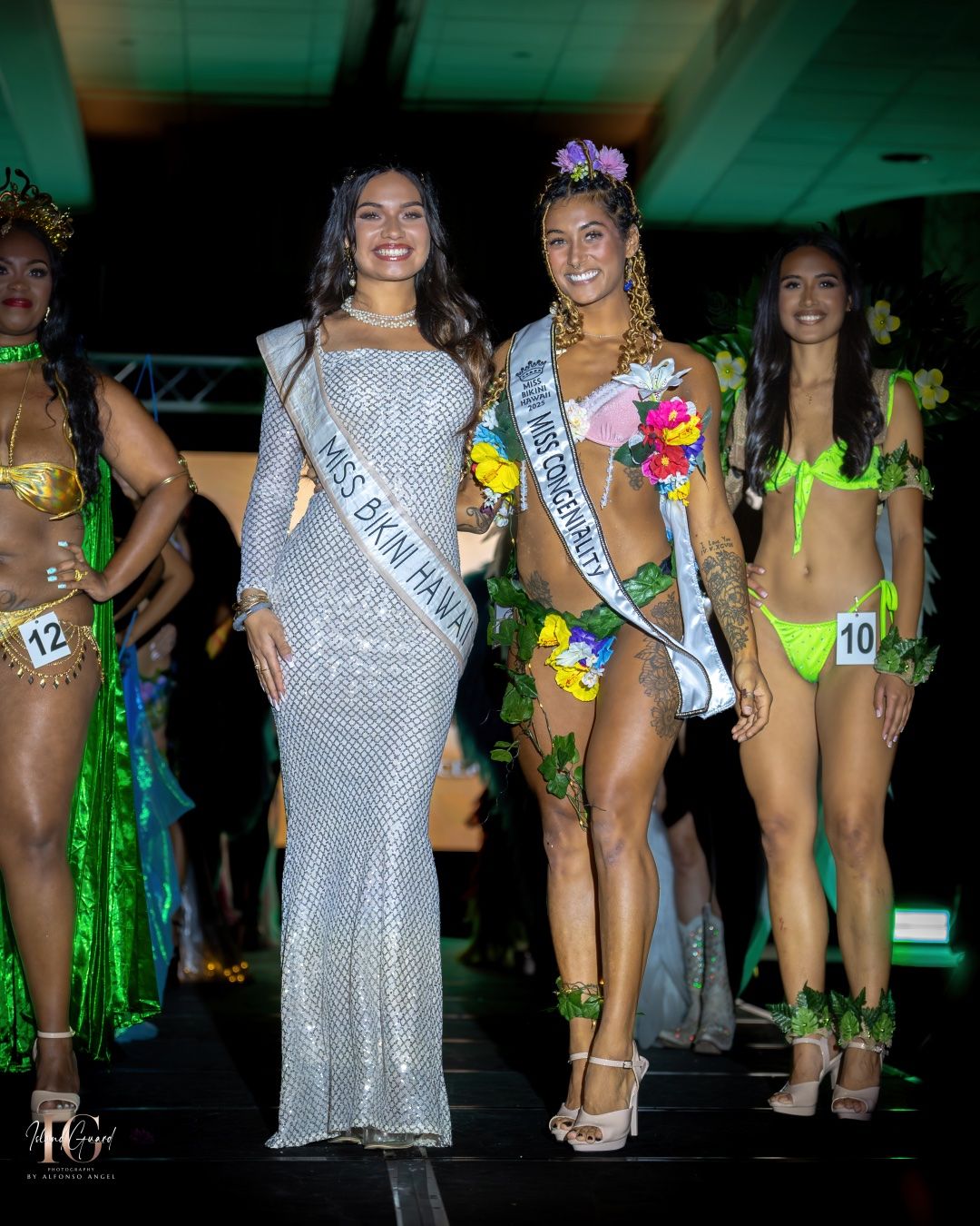 Beauty pageant contestants pose together on stage wearing evening gowns and tropical costumes under green lighting.