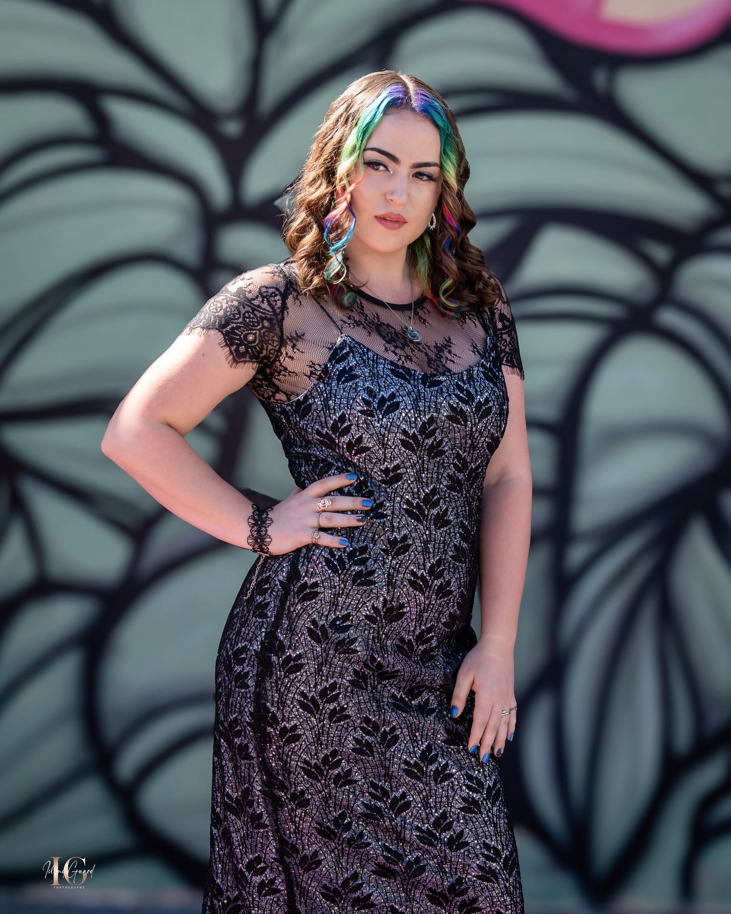 Fashion model wearing black lace dress poses against artistic wall mural backdrop.