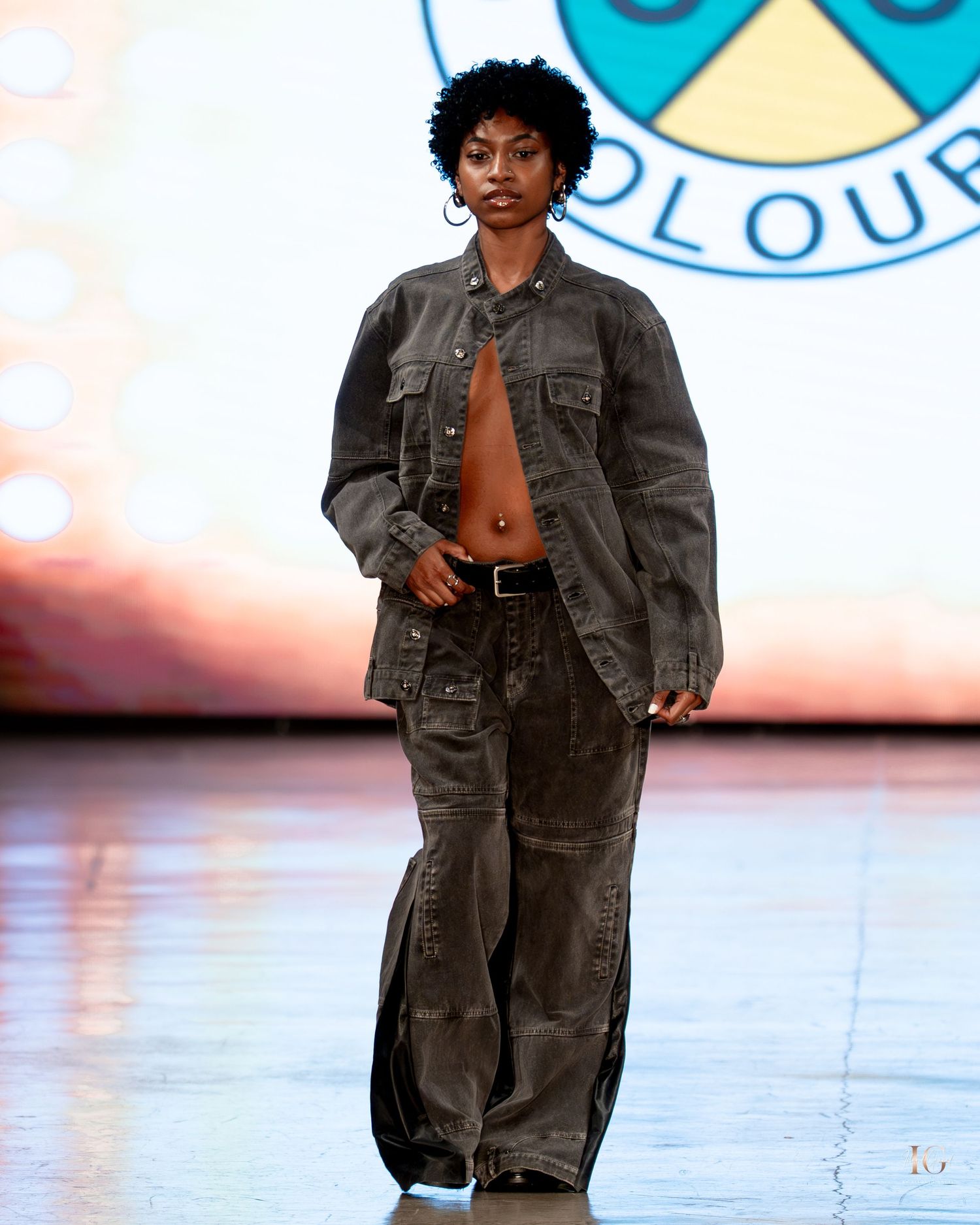 Model walks runway in dark denim ensemble during fashion show with water-like reflective floor.