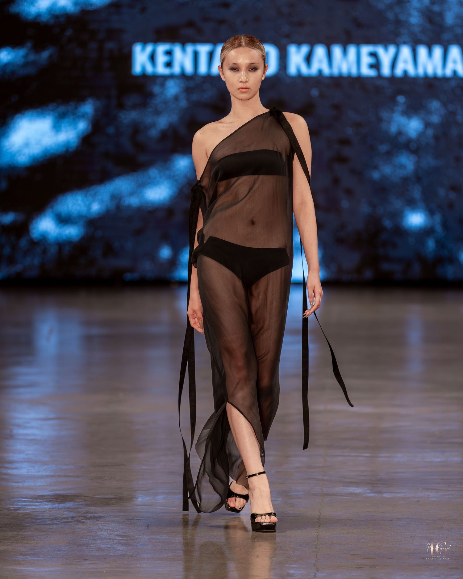 Model walks runway in sheer black asymmetrical dress during fashion show with dramatic blue lighting.