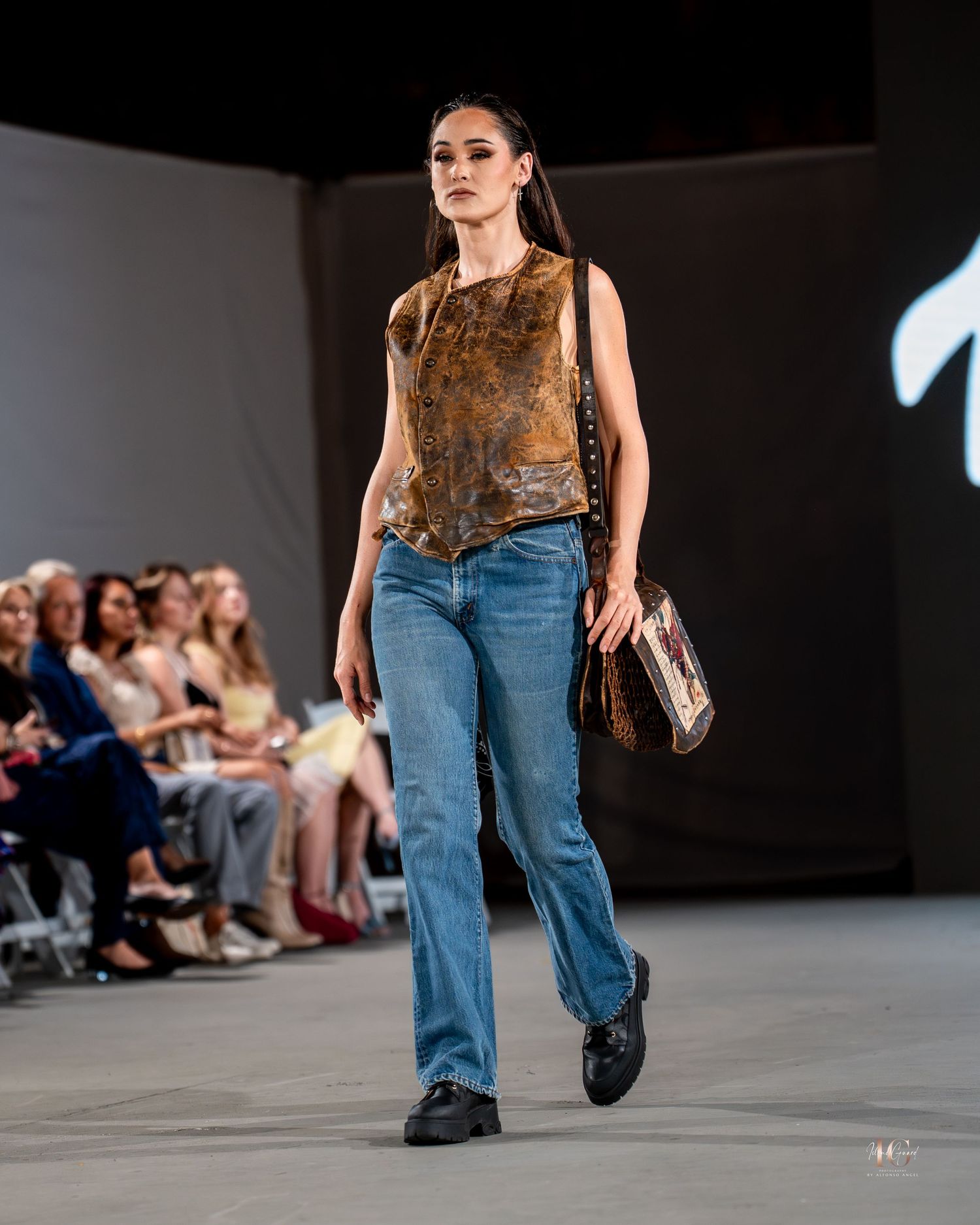 Model walks runway in brown fur vest with blue jeans and black boots during fashion show.