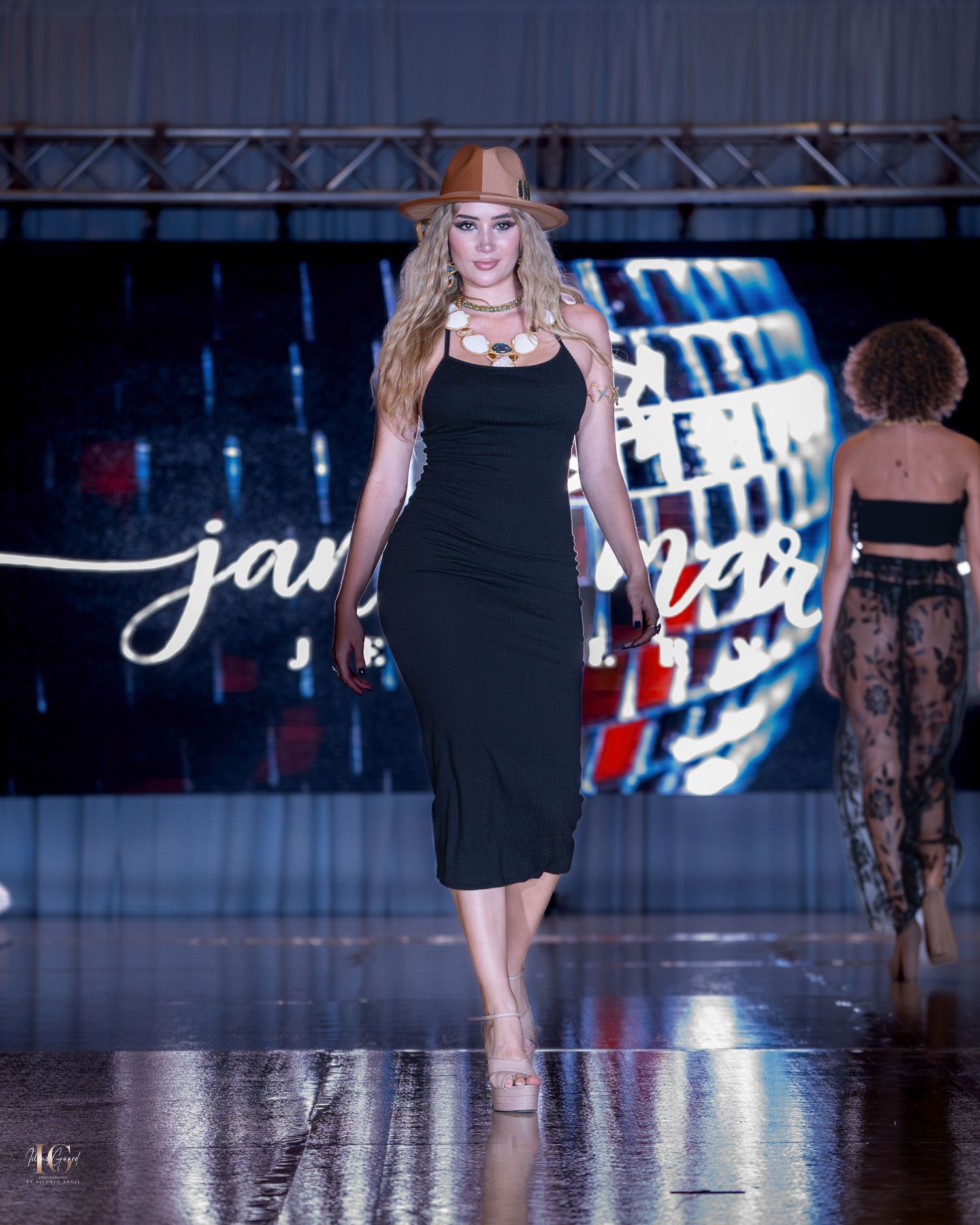 Models walk the runway in elegant black dresses during a fashion show with dramatic blue lighting and reflective floors.