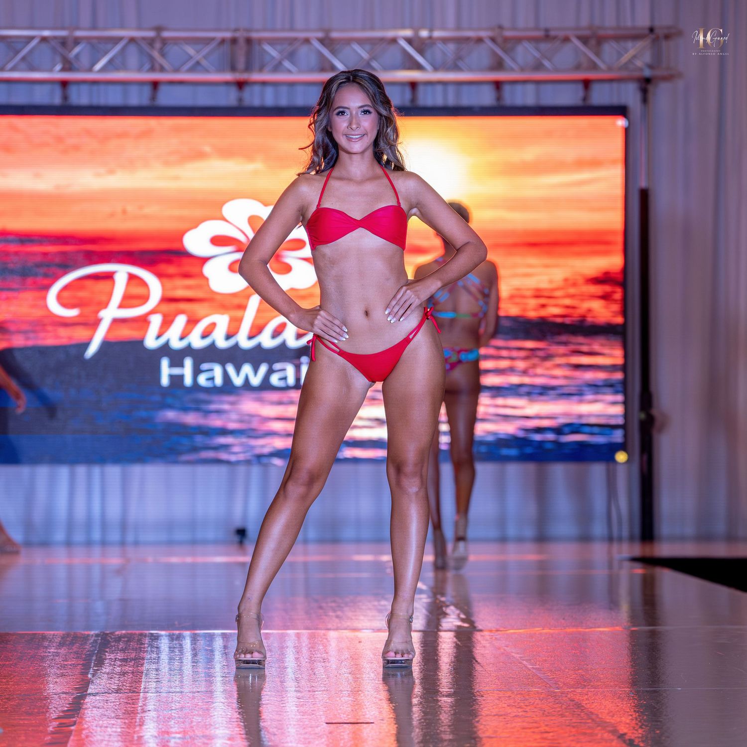 Models walk the runway in red swimwear at a beachwear fashion show with sunset backdrop lighting.