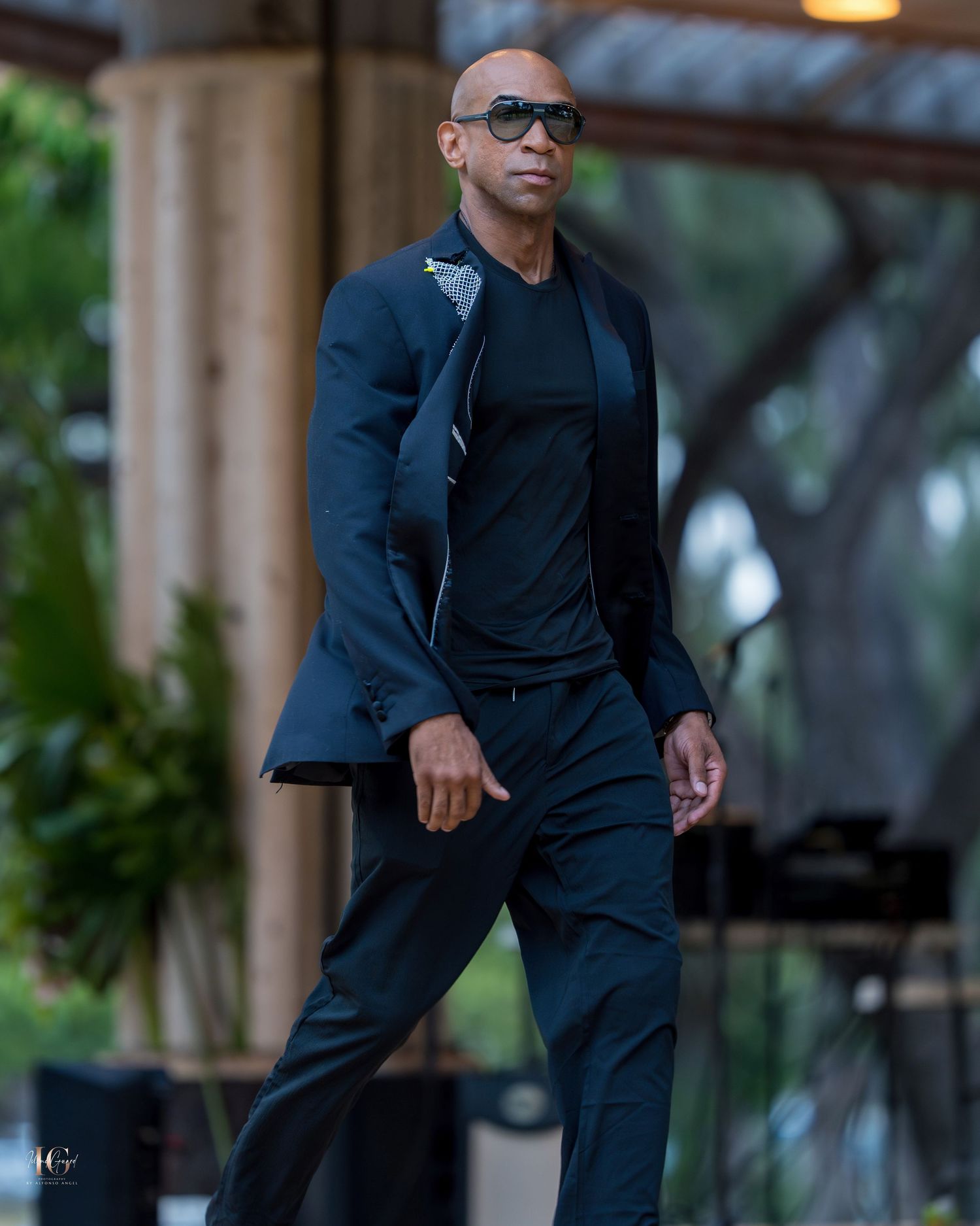 Someone wearing a dark suit and glasses stands in a contemplative pose against a blurred outdoor background.