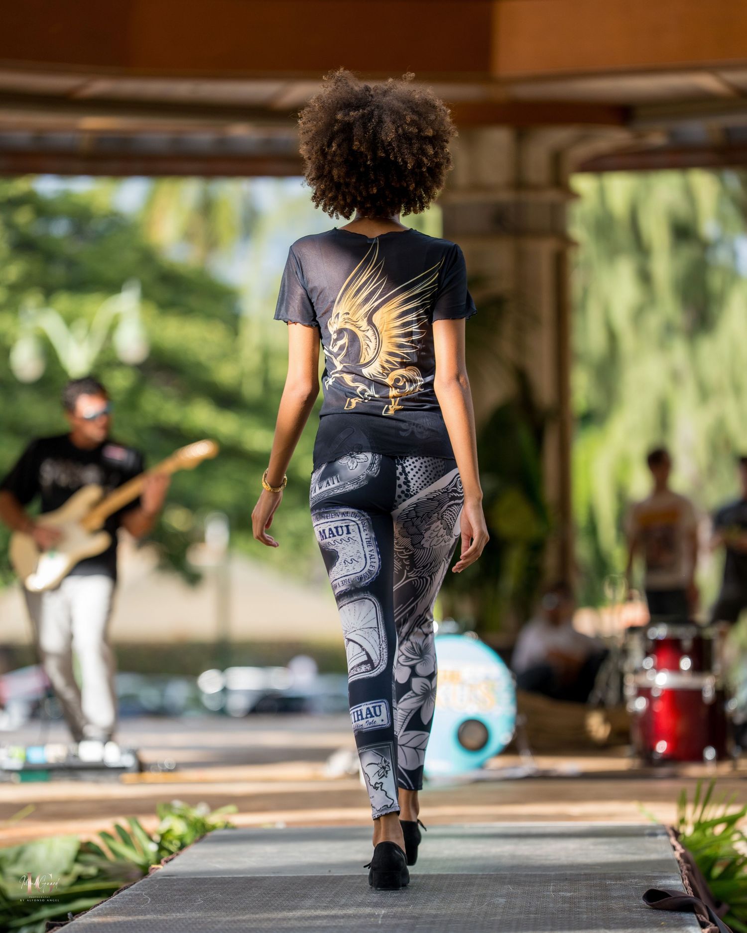 A person wearing patterned pants walks on a stage with musicians performing in the background at an outdoor venue.