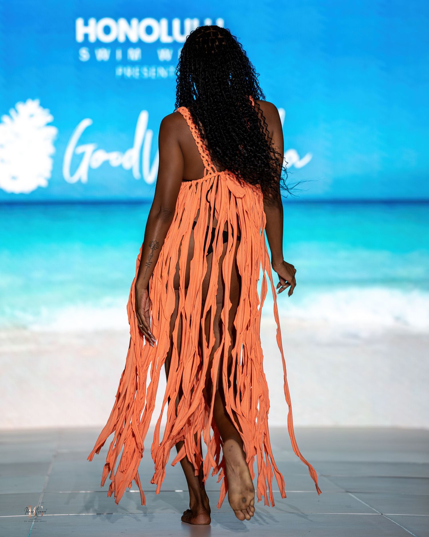 Back view of coral fringe swimwear dress against beach backdrop at fashion show.
