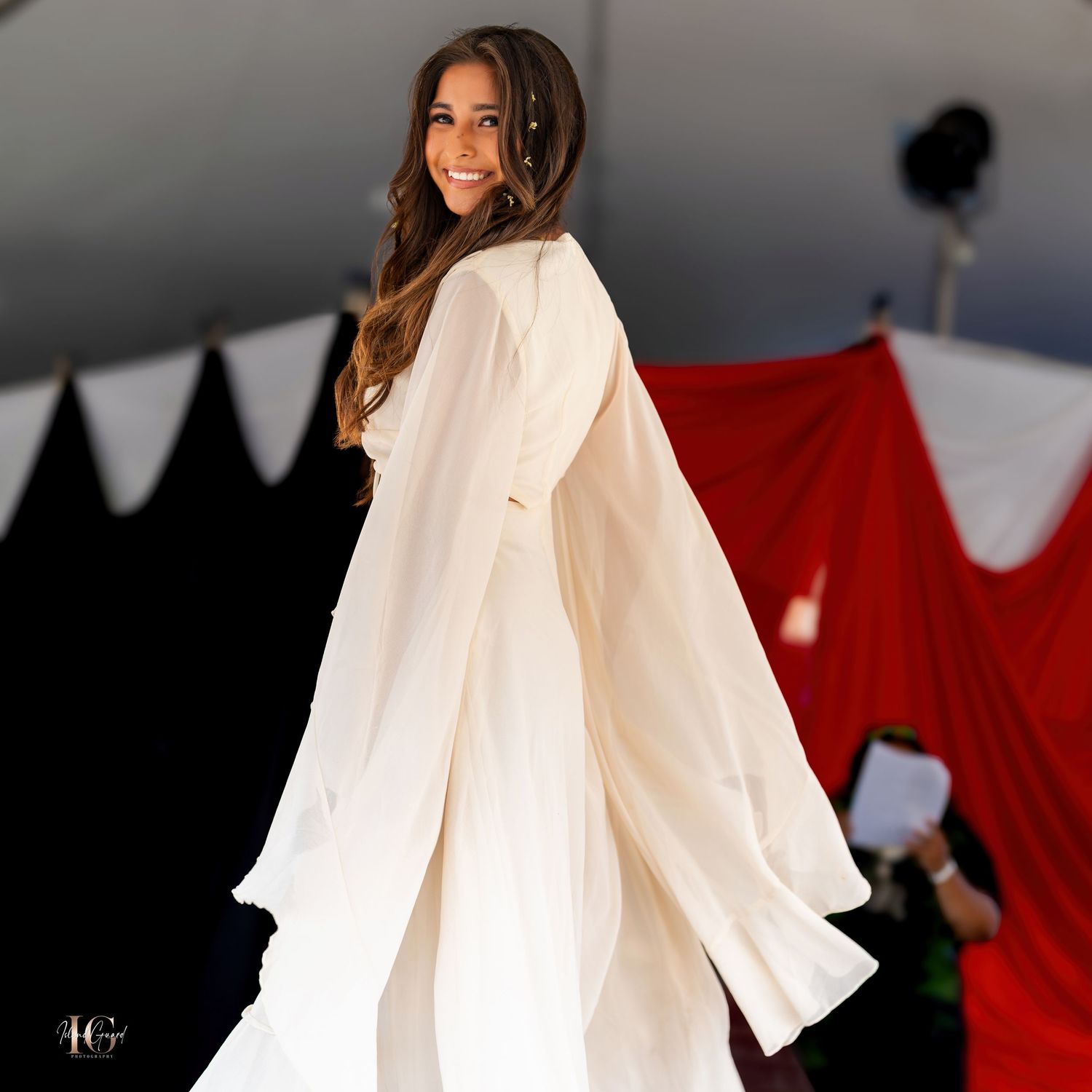 Model wearing ethereal white flowing sleeve top poses against red backdrop.