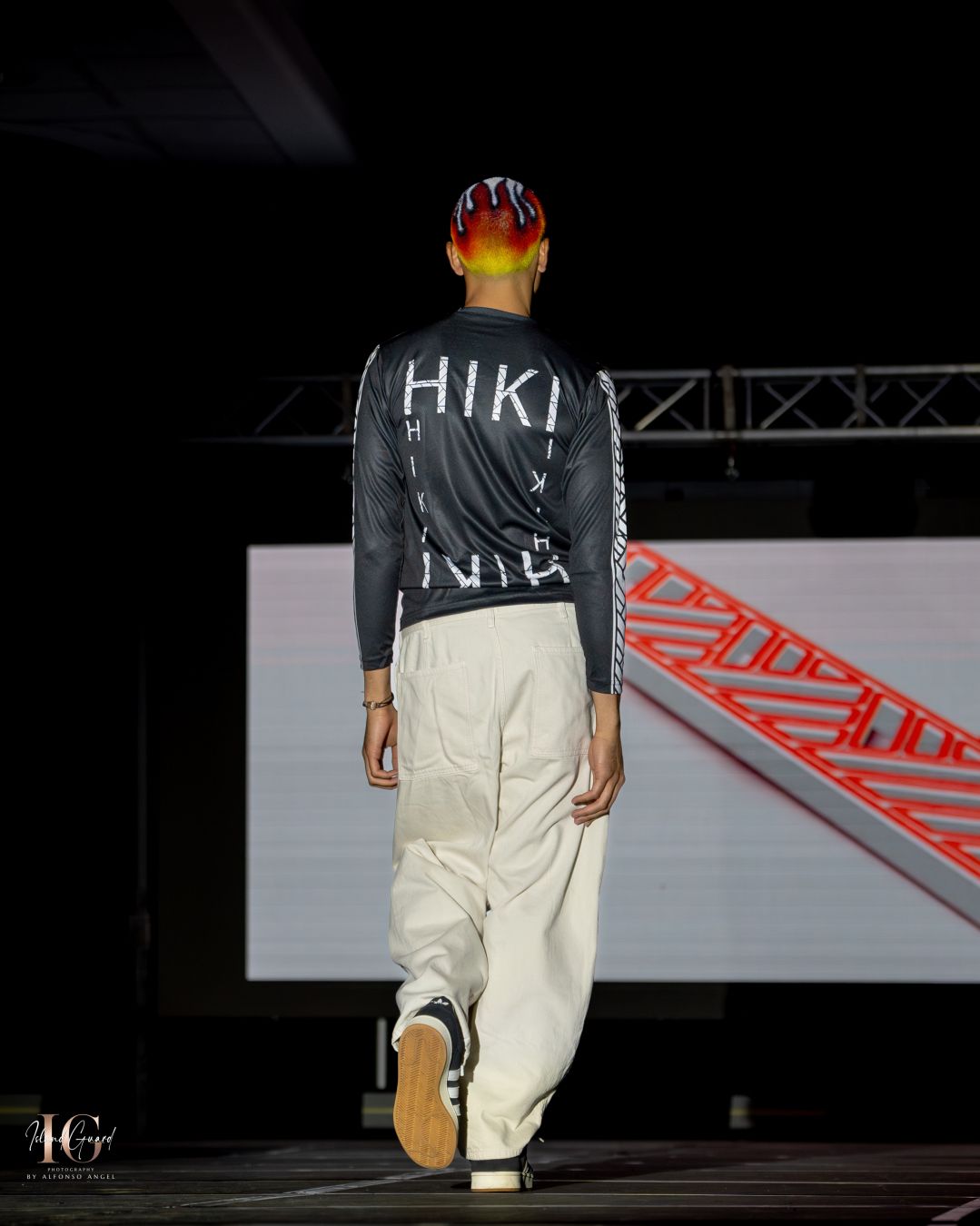 Model walks runway in black shirt and white pants at fashion show with red signage in background.