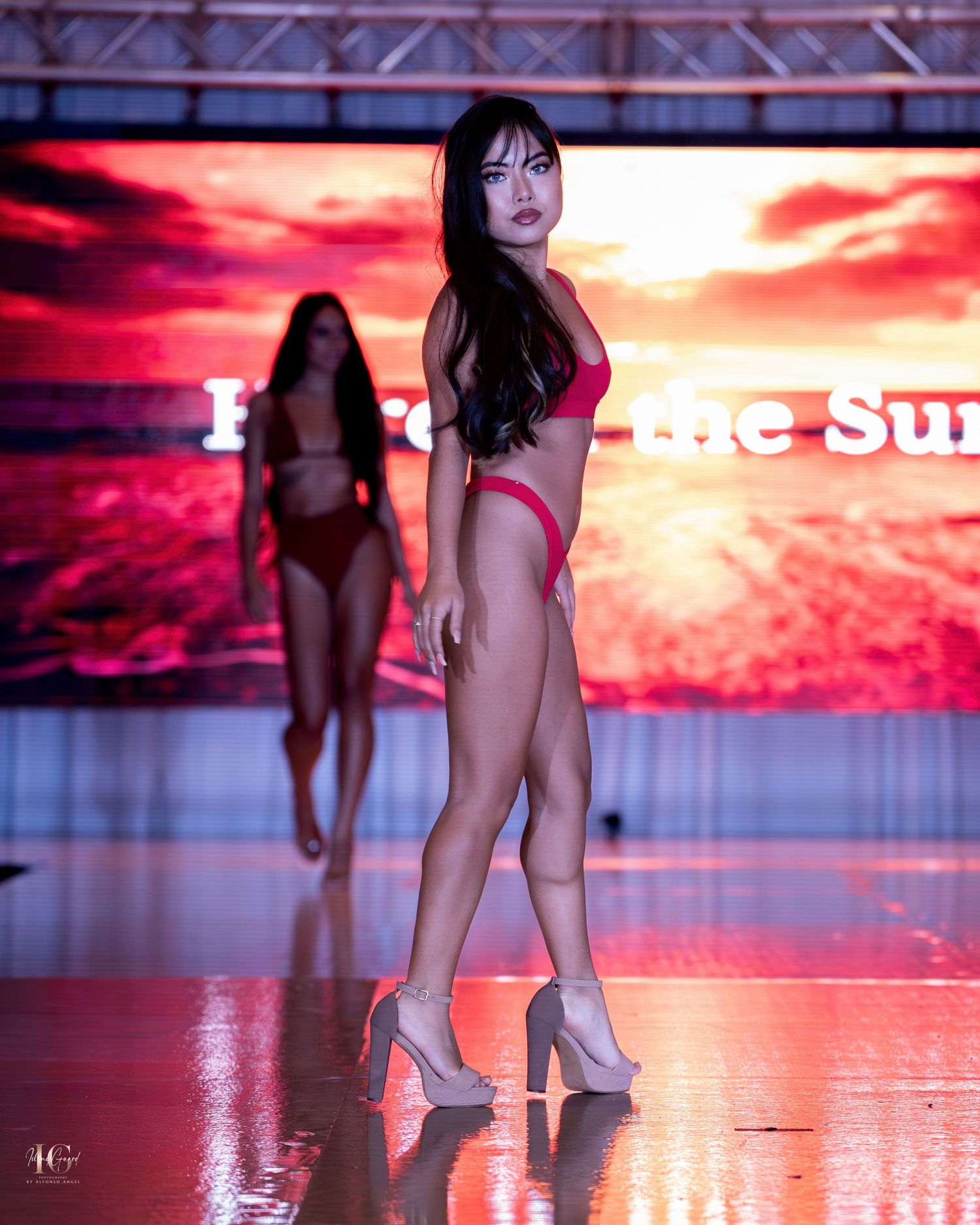 Model in red swimwear strikes pose on runway against sunset backdrop display.