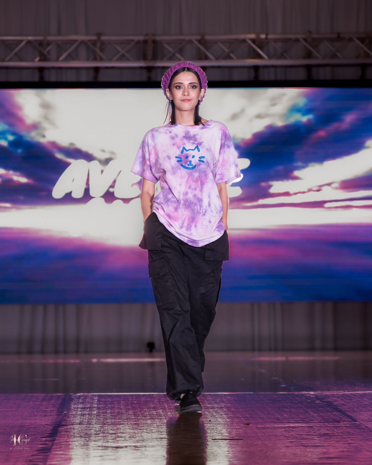 Model walks runway in tie-dye shirt and dark pants against purple-lit backdrop.