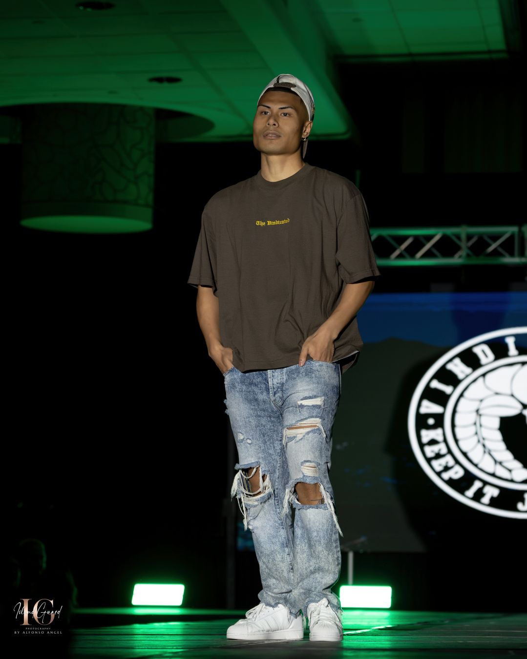 Model wearing casual streetwear with ripped jeans and baseball cap poses on illuminated runway stage.