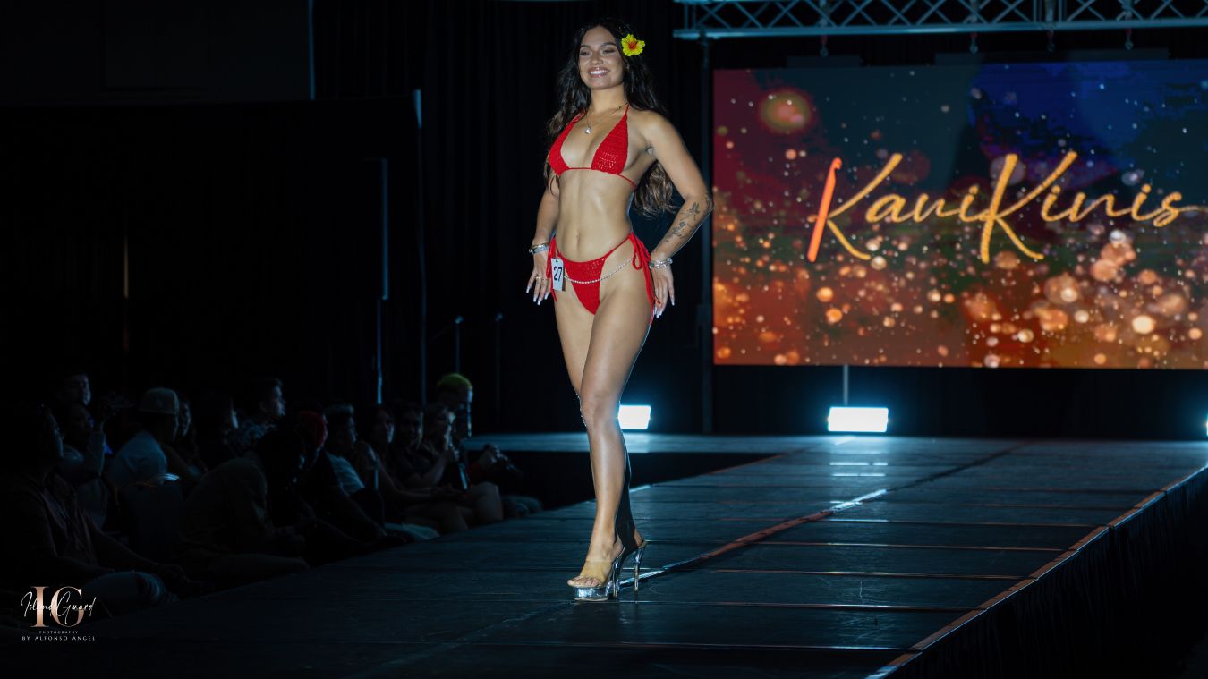 Model walks runway in red swimwear against sparkly backdrop.