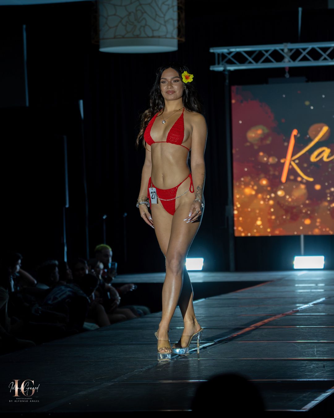 Model showcases red bikini on catwalk with glowing effects in background.