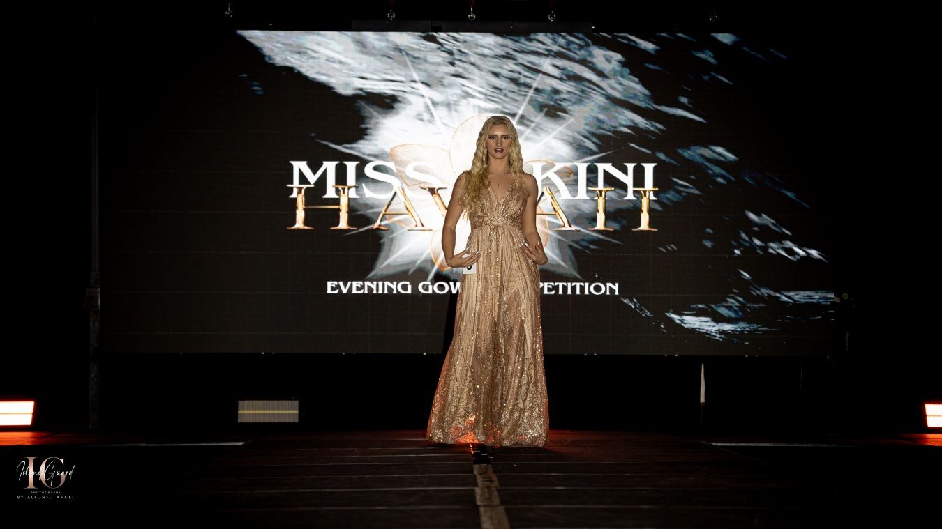 A person in a flowing gold dress stands on stage with dramatic lighting and projected backdrop effects during a performance.