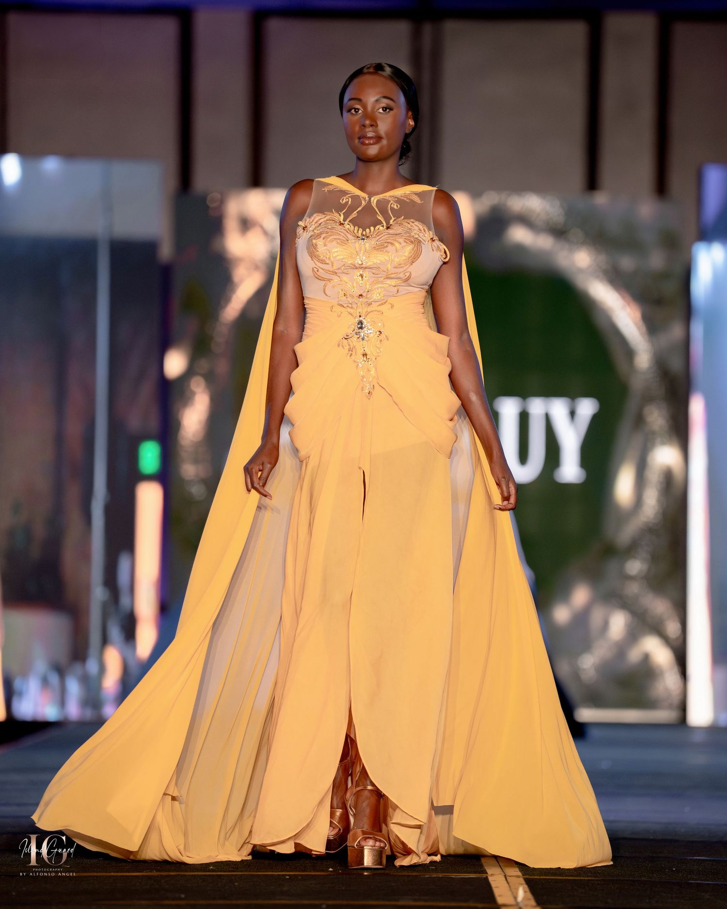 Model wearing elegant yellow cape gown with embellished bodice on fashion runway.