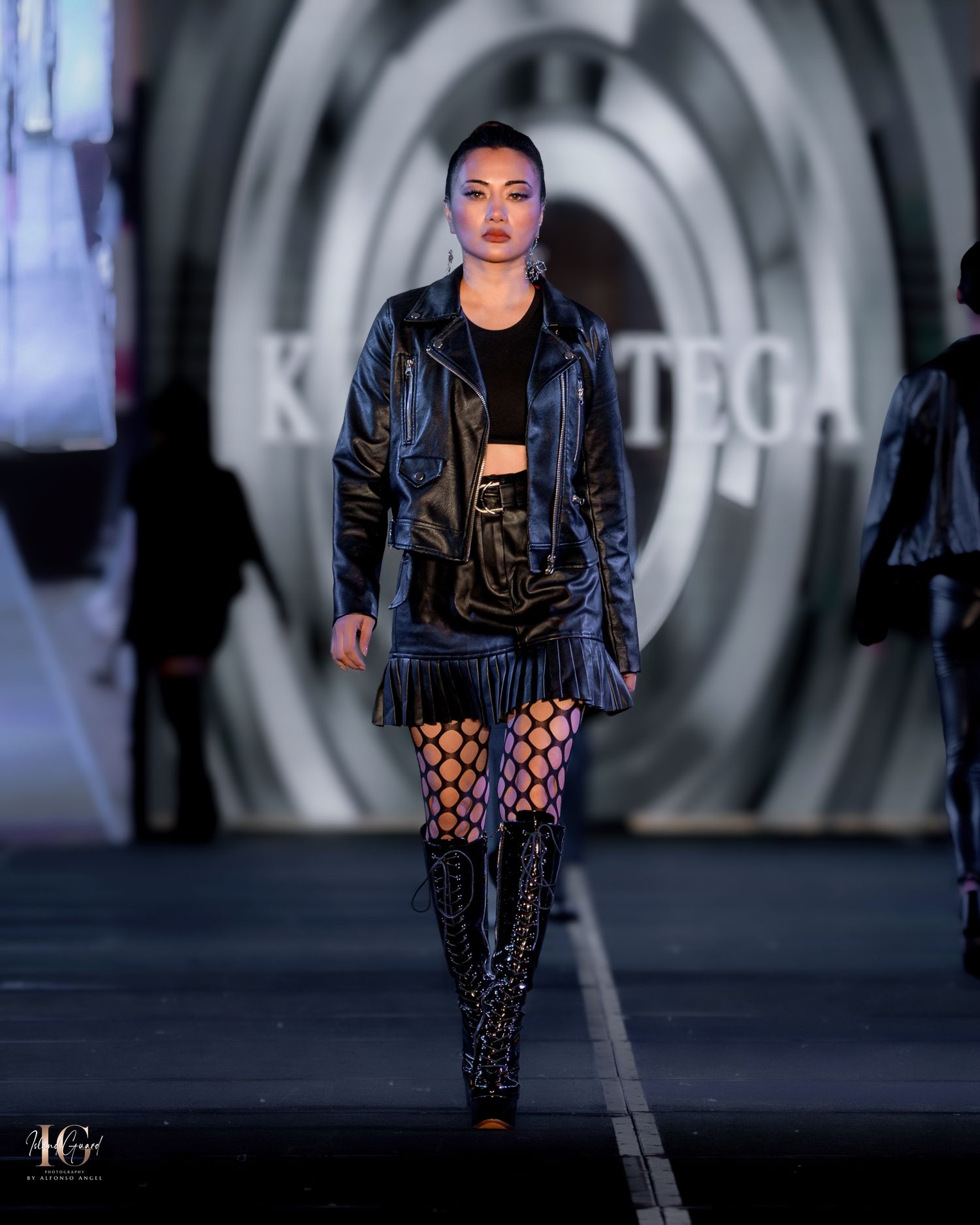 Model wears edgy black leather jacket with fishnet stockings and combat boots on runway.