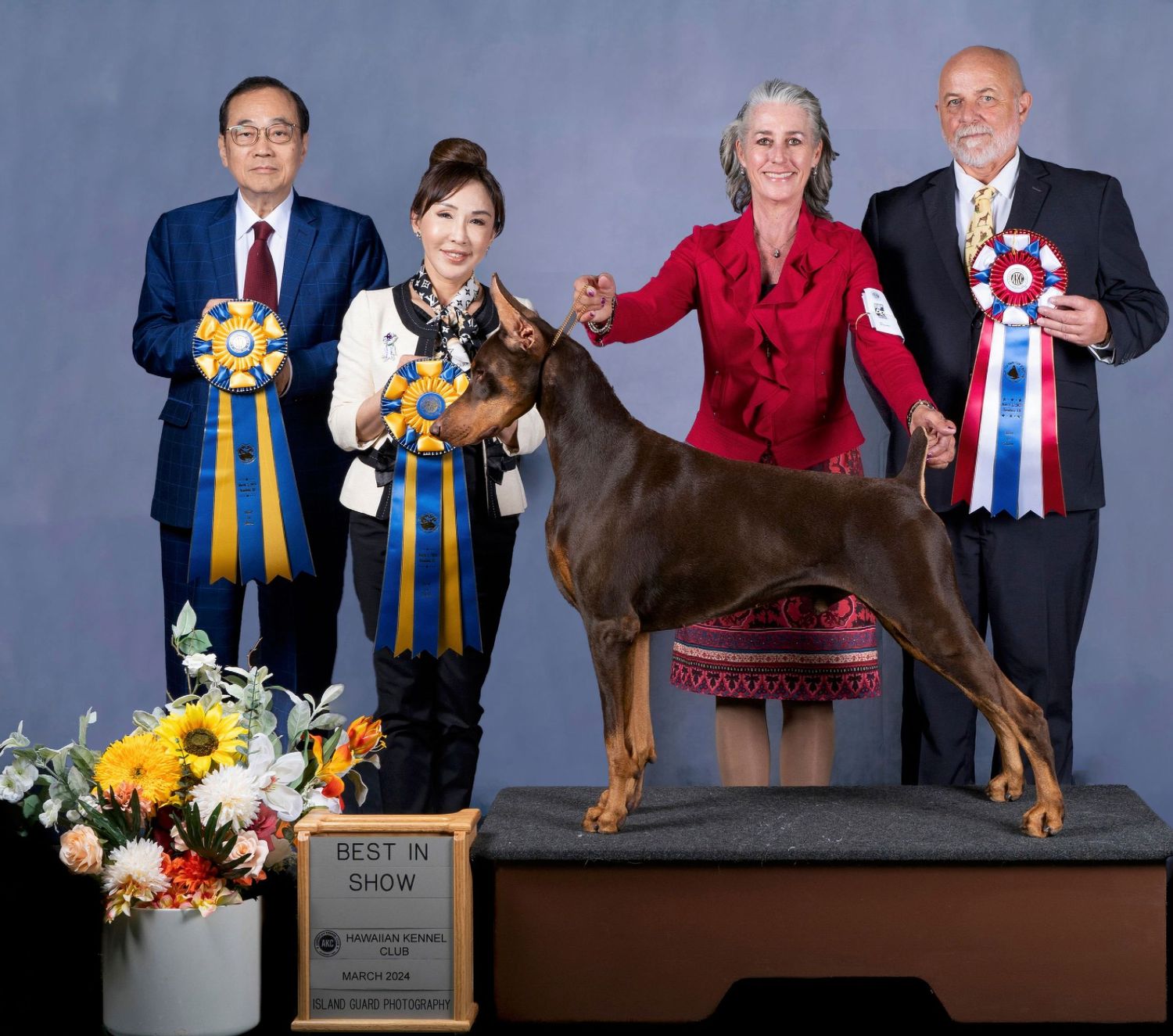 Four people proudly display ribbons at a Best in Show dog competition with a Doberman Pinscher on a podium.