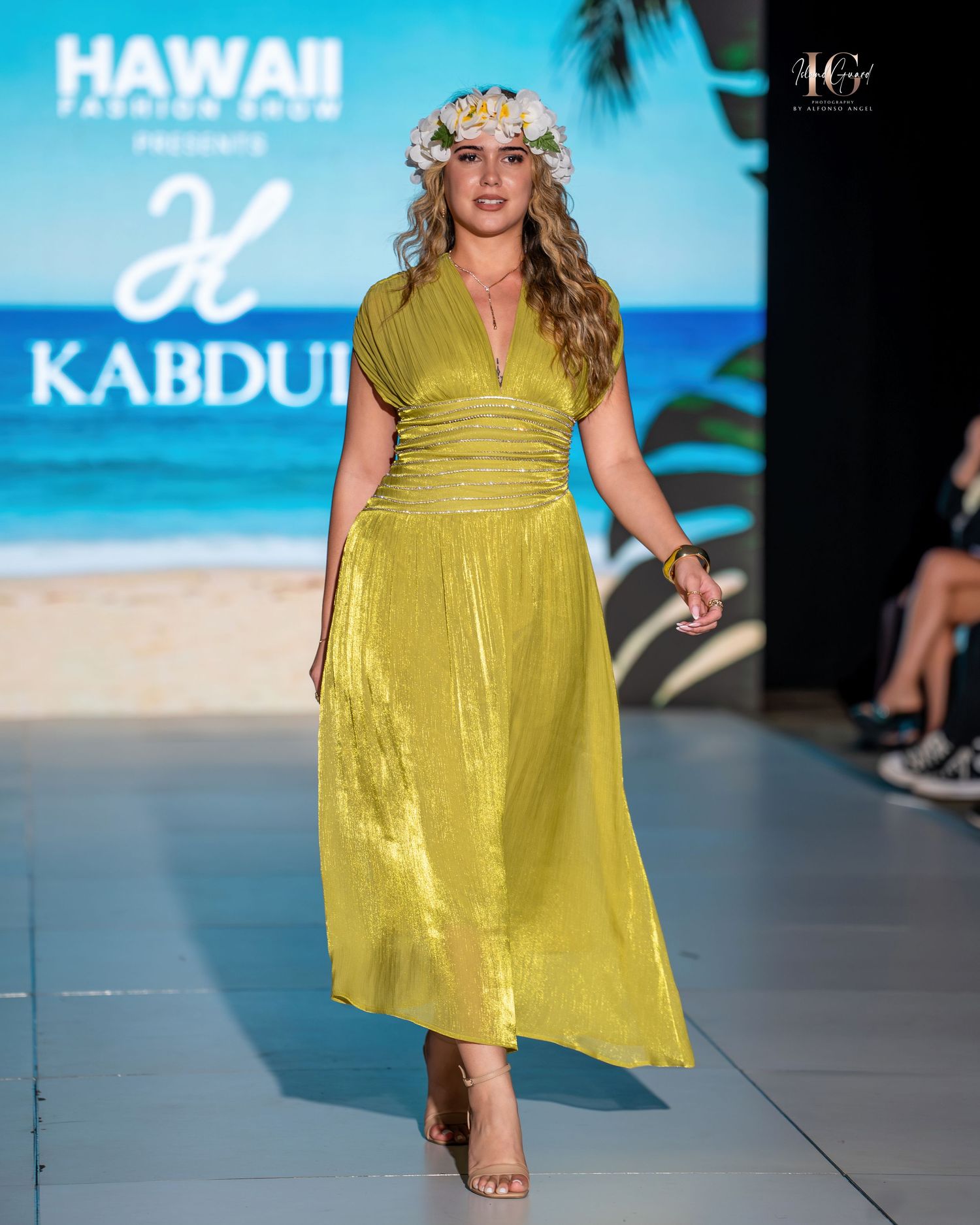 Model walks runway in flowy yellow sundress at tropical-themed fashion show with palm trees and beach backdrop.