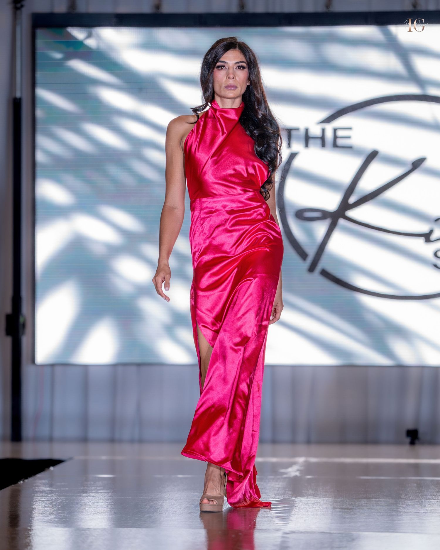 Model walks runway in vibrant hot pink satin dress with asymmetrical draping at fashion show.