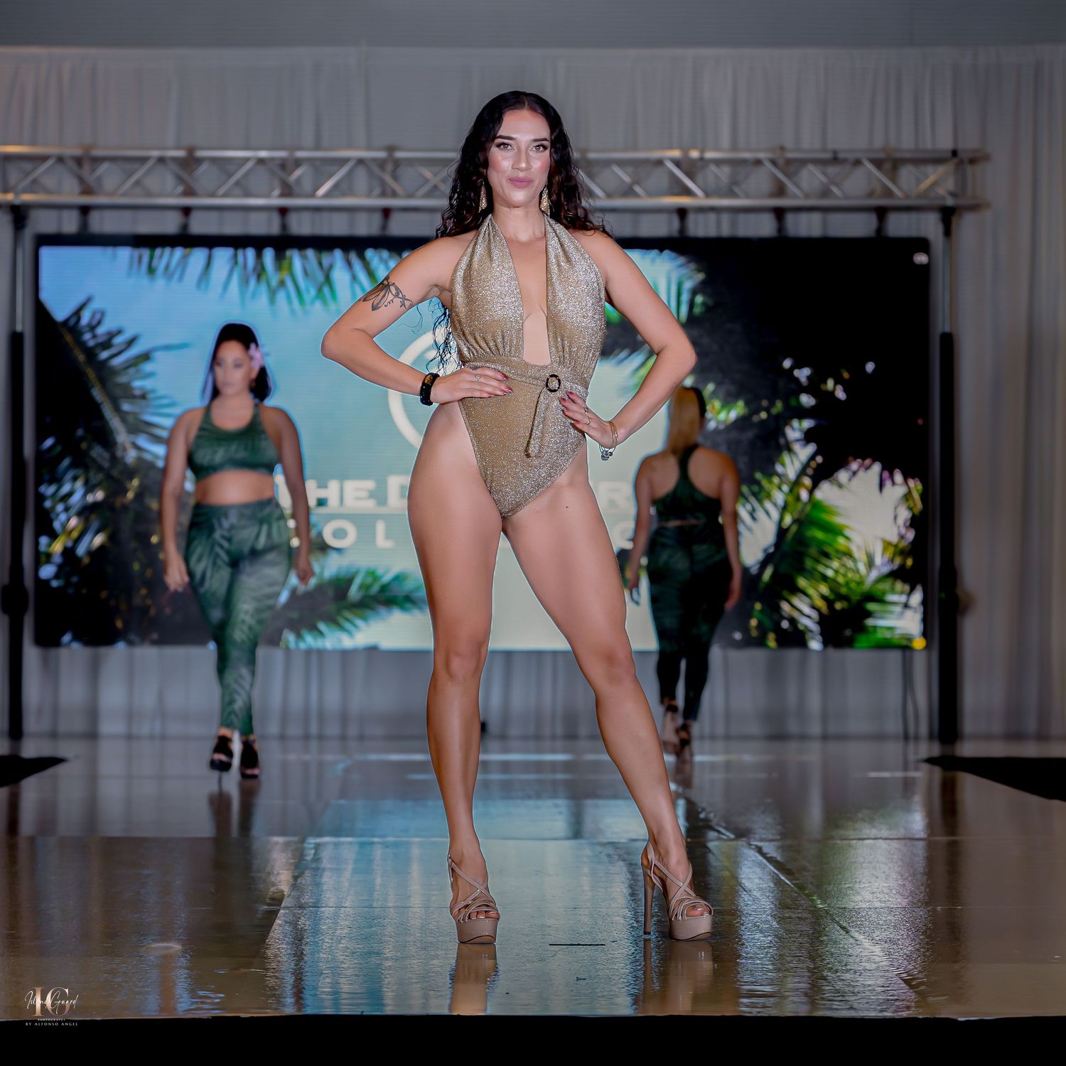 Models walk the runway wearing swimwear at a fashion show with palm leaf decorations in the background.