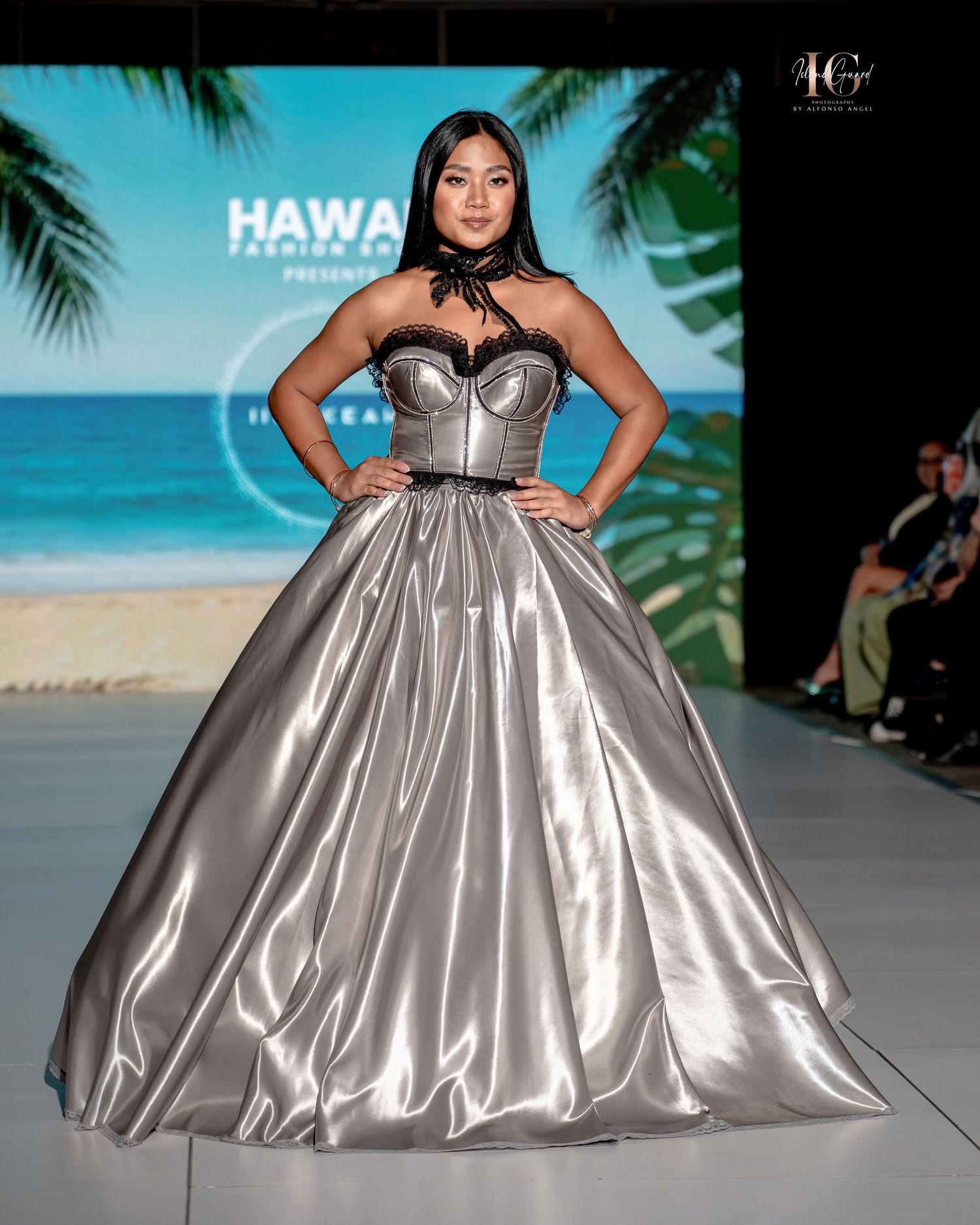Model walks runway in shimmering silver ballgown with sweetheart neckline at tropical beach-themed fashion show.